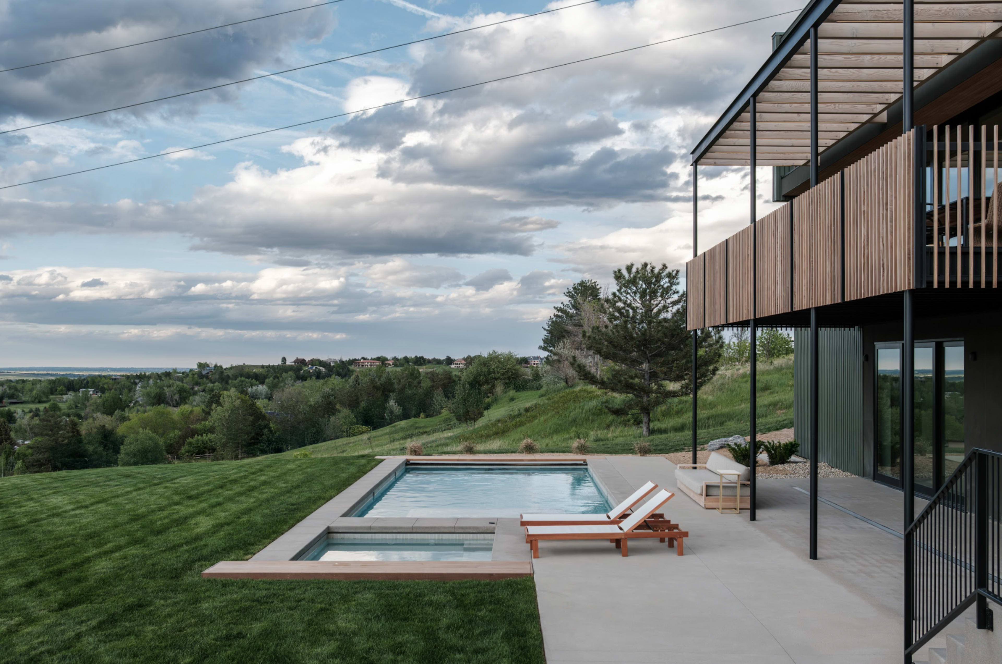 A modern house with a wooden exterior overlooks a landscaped yard featuring a swimming pool and lounge chairs under a cloudy sky.