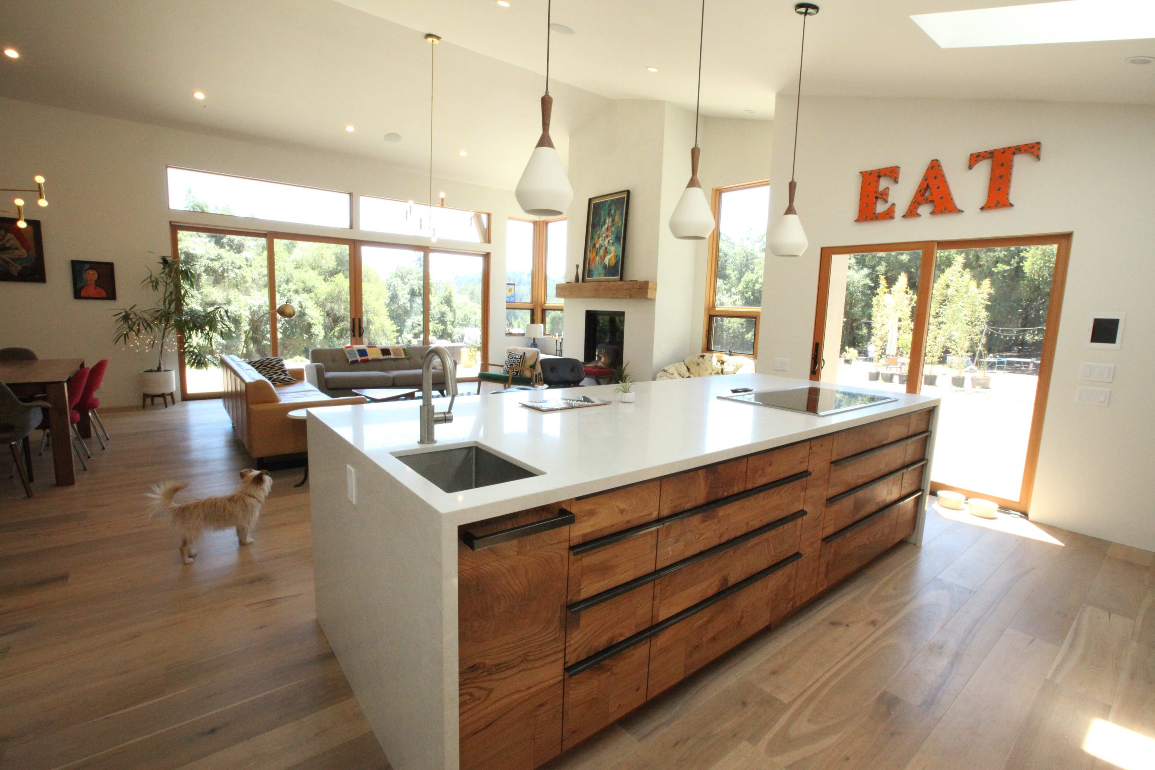 A modern kitchen features a large white island with wooden cabinets and a living area visible in the background.