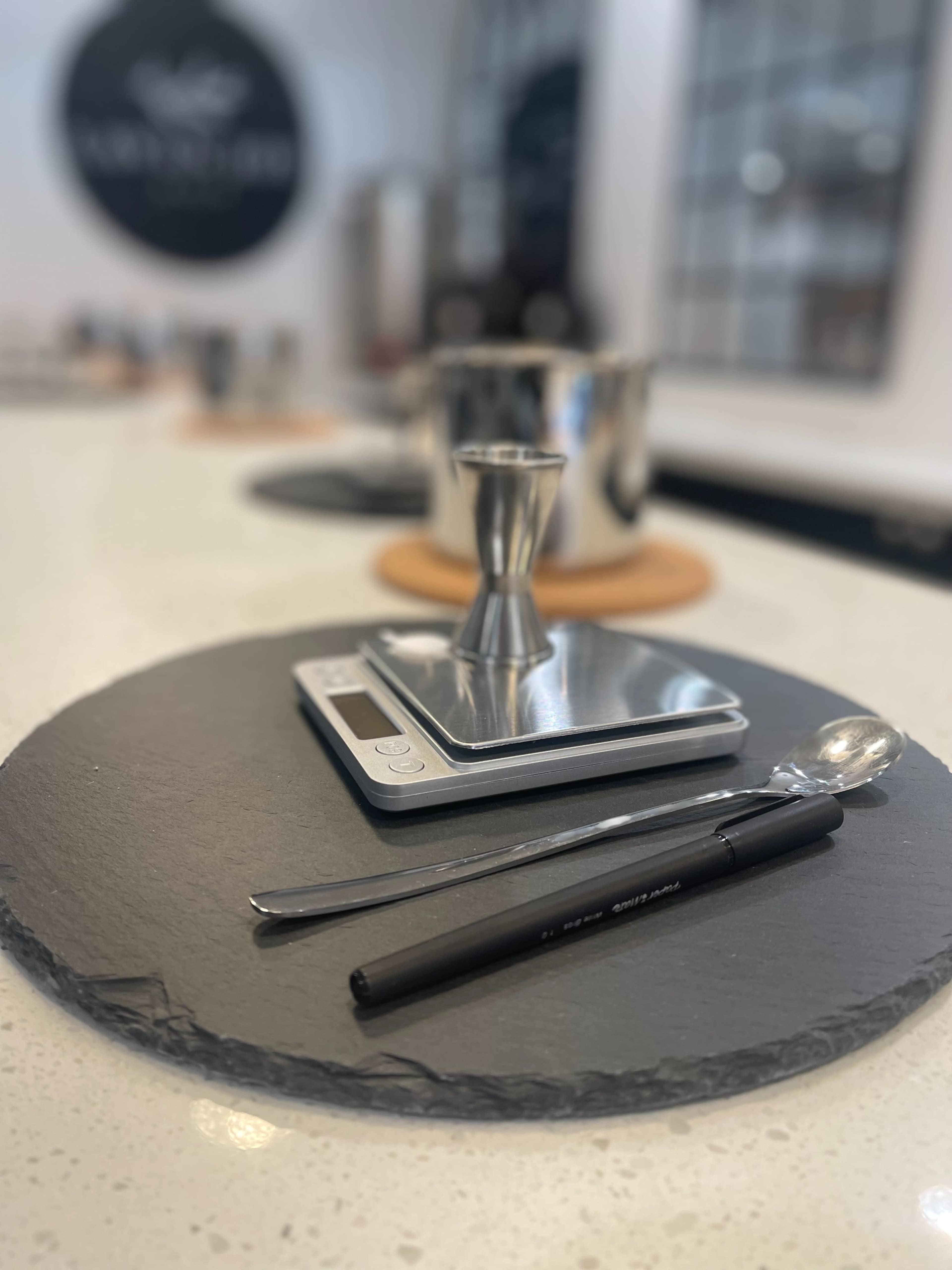 A digital scale, a jigger, a spoon, and a pen are placed on a round slate surface in a kitchen setting.