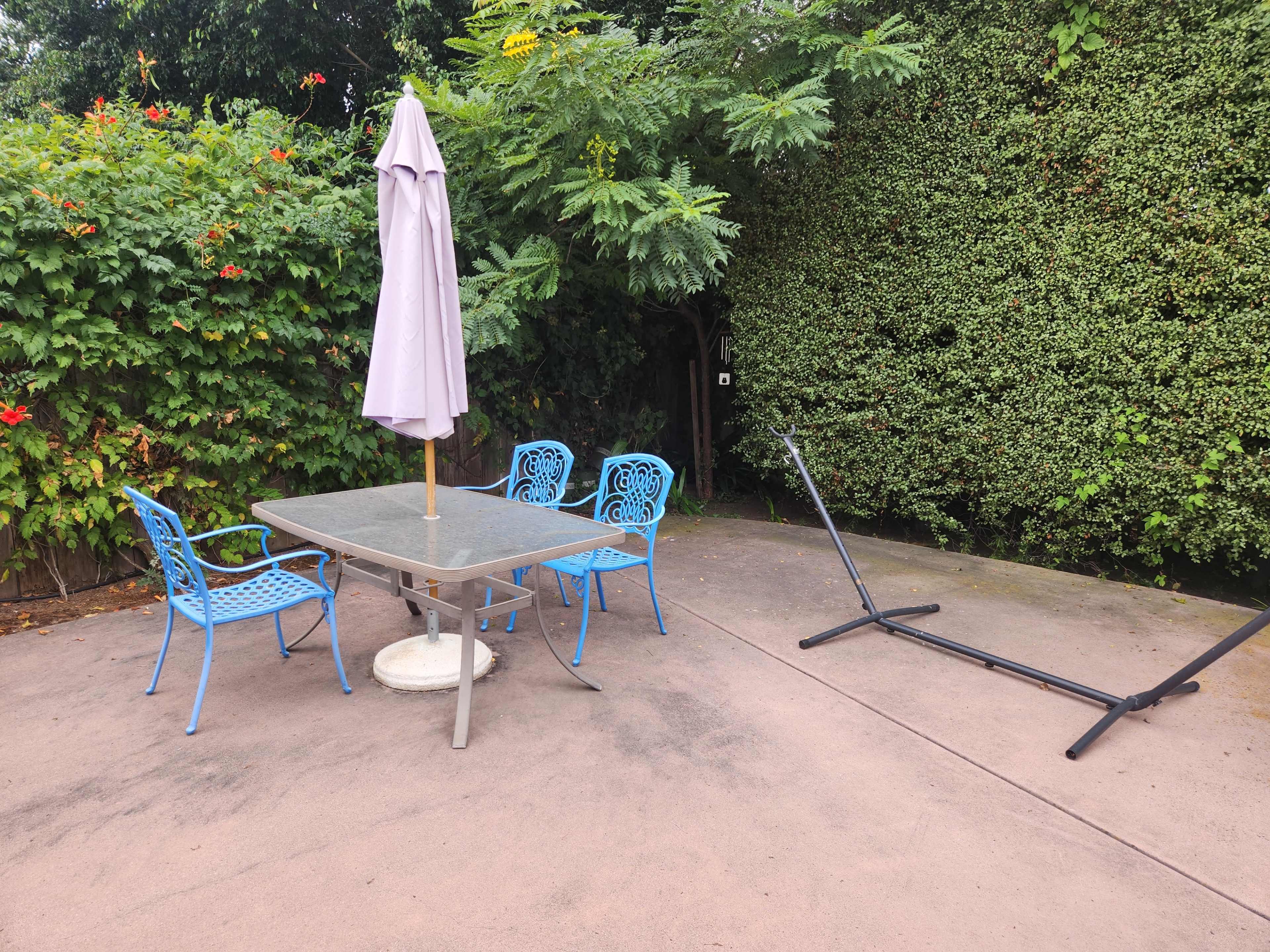 A patio area features a table with four blue chairs and an umbrella, alongside a disassembled hammock stand amidst greenery.