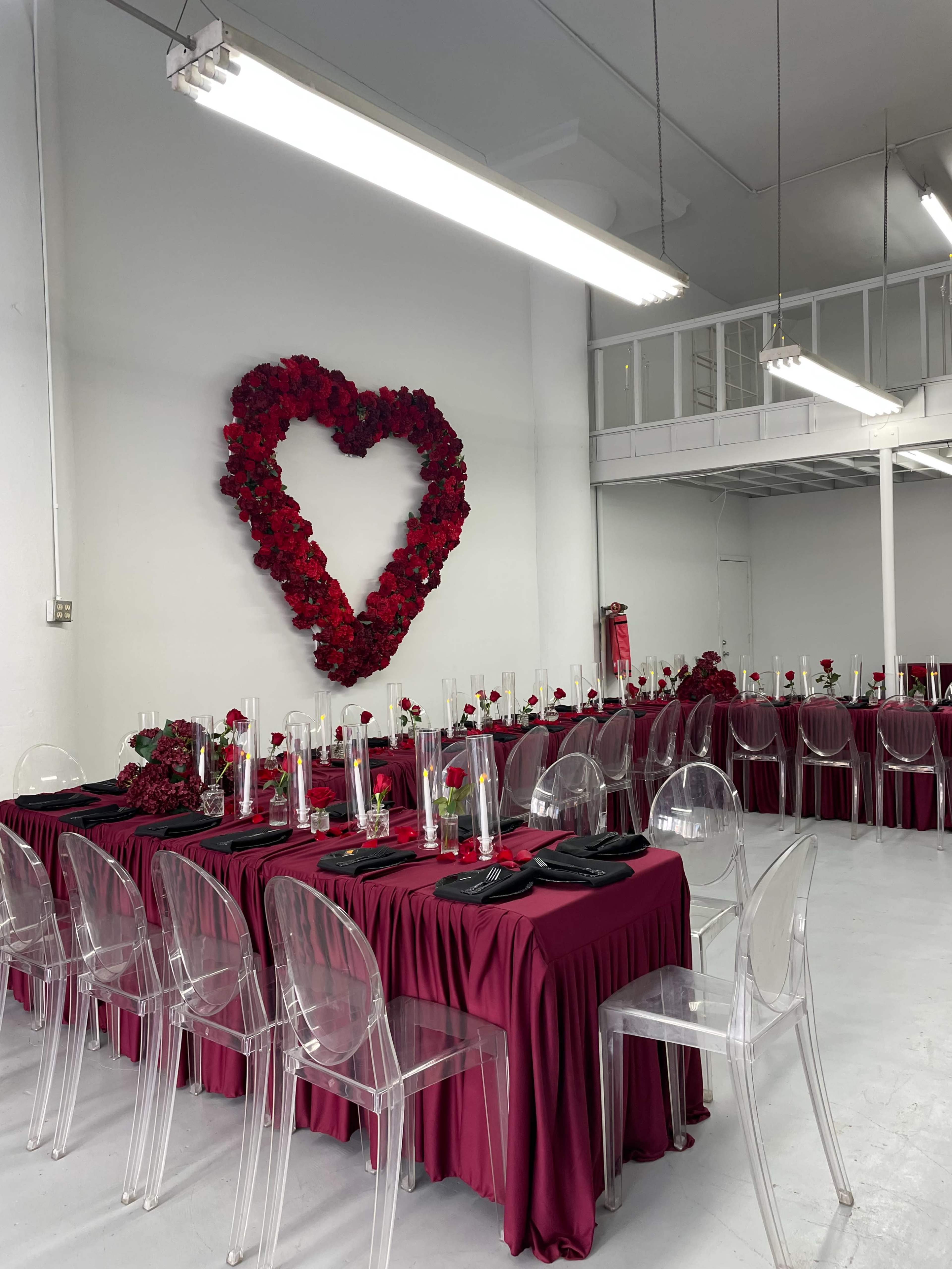 The image shows a banquet hall set up for an event, featuring tables adorned with red tablecloths, clear chairs, and a heart-shaped arrangement of red flowers on the wall.