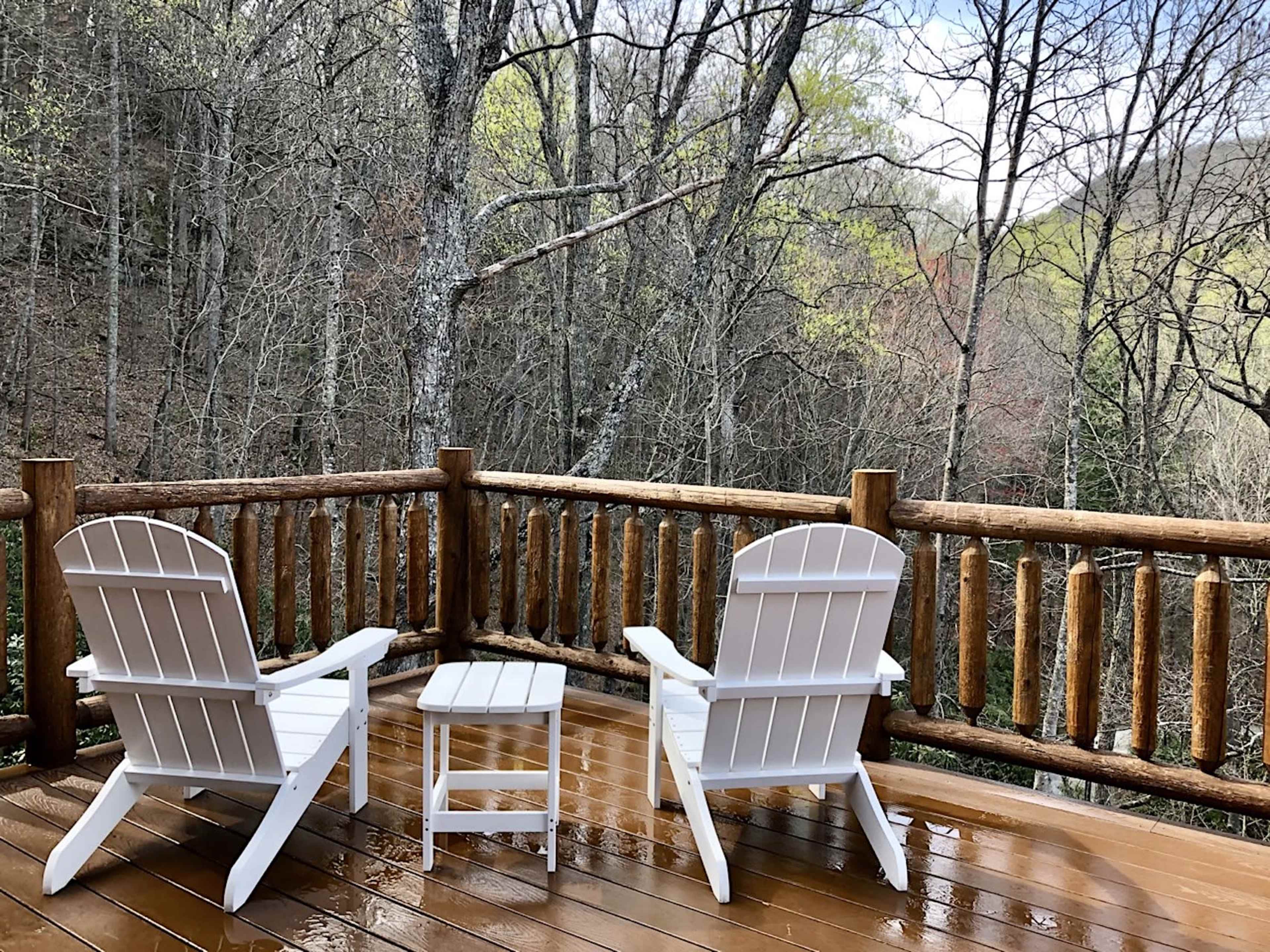 Two white Adirondack chairs and a small table sit on a wooden deck overlooking a forested landscape.