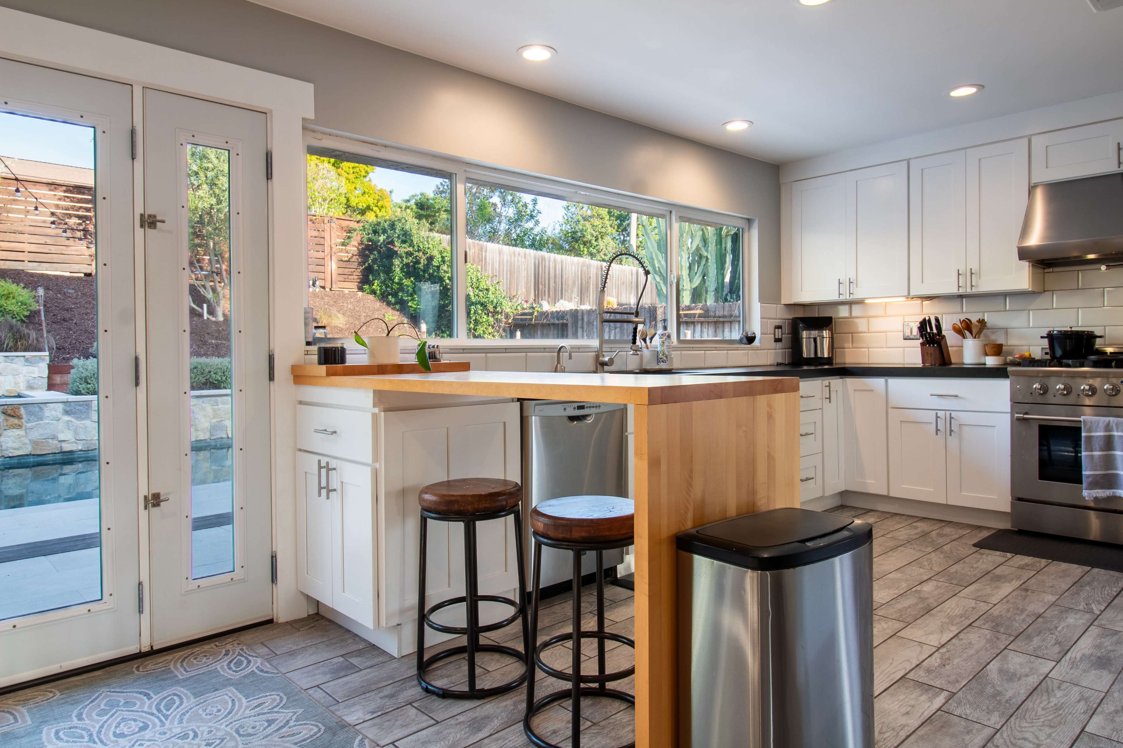 A modern kitchen features a central island with stools, stainless steel appliances, and large windows overlooking a backyard pool area.