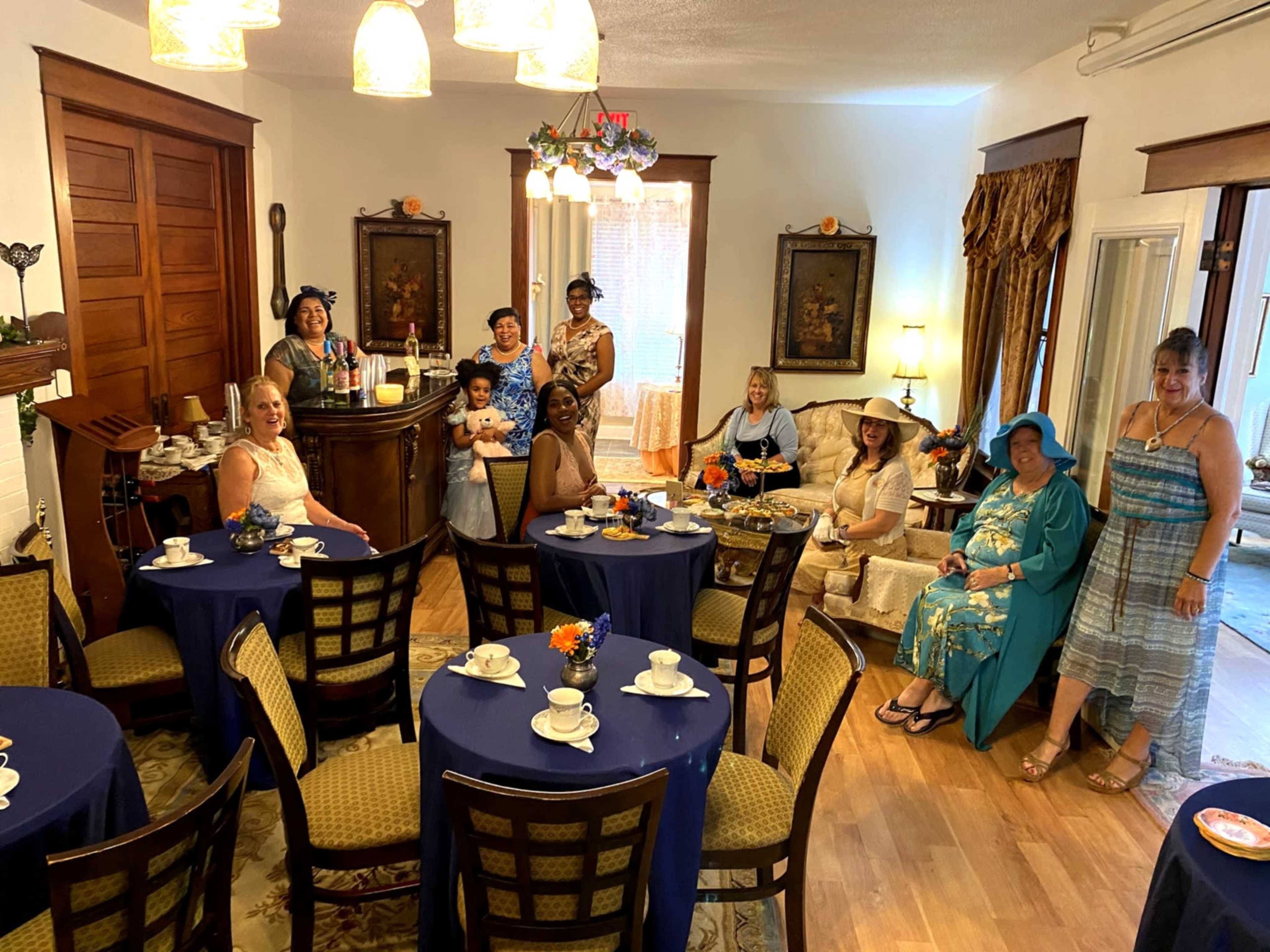 A group of women in formal attire pose together in a warmly decorated tea room with tables set for tea.
