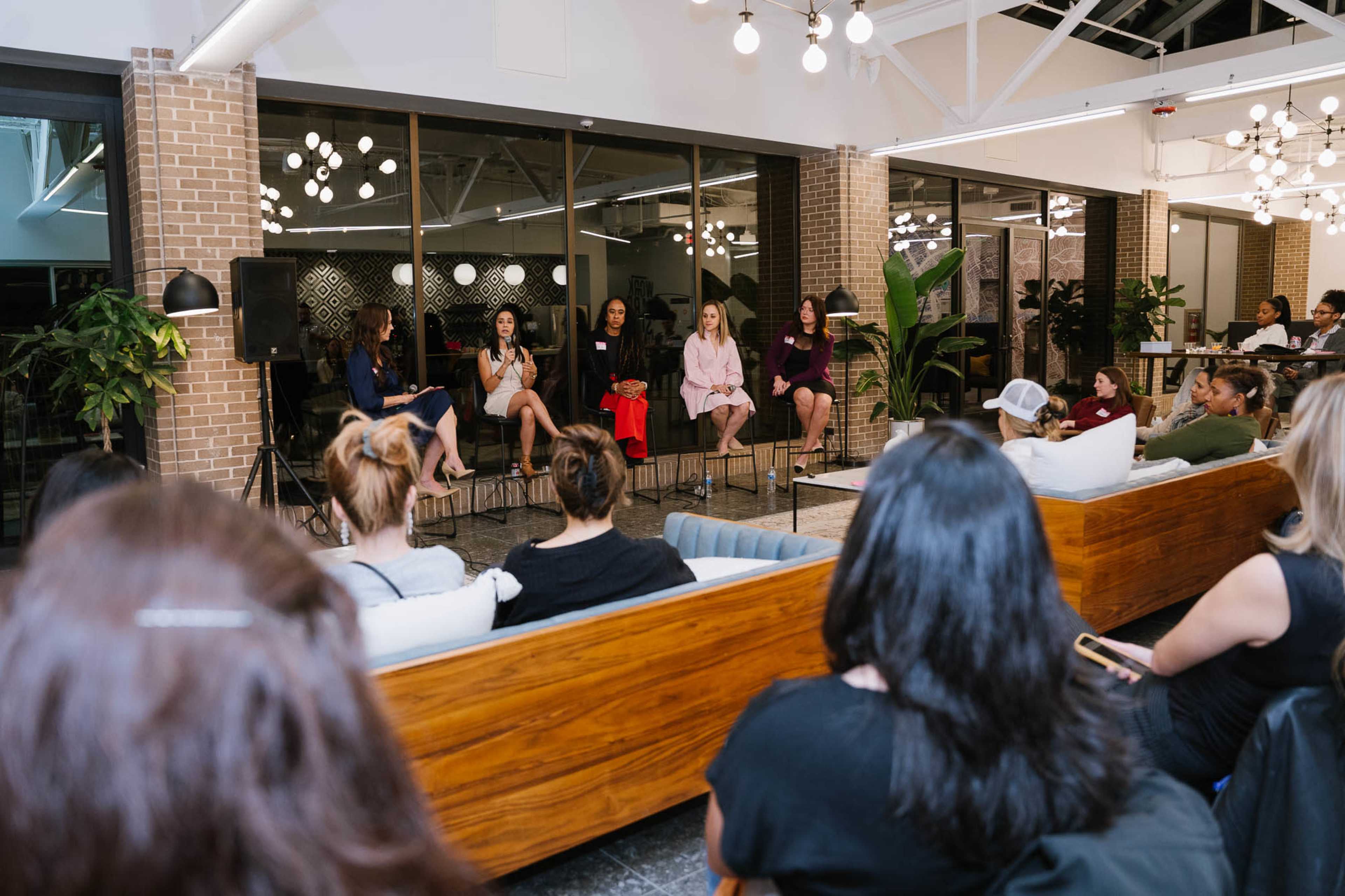 A panel of five women speaks to an audience in a modern indoor space with seating and plants.