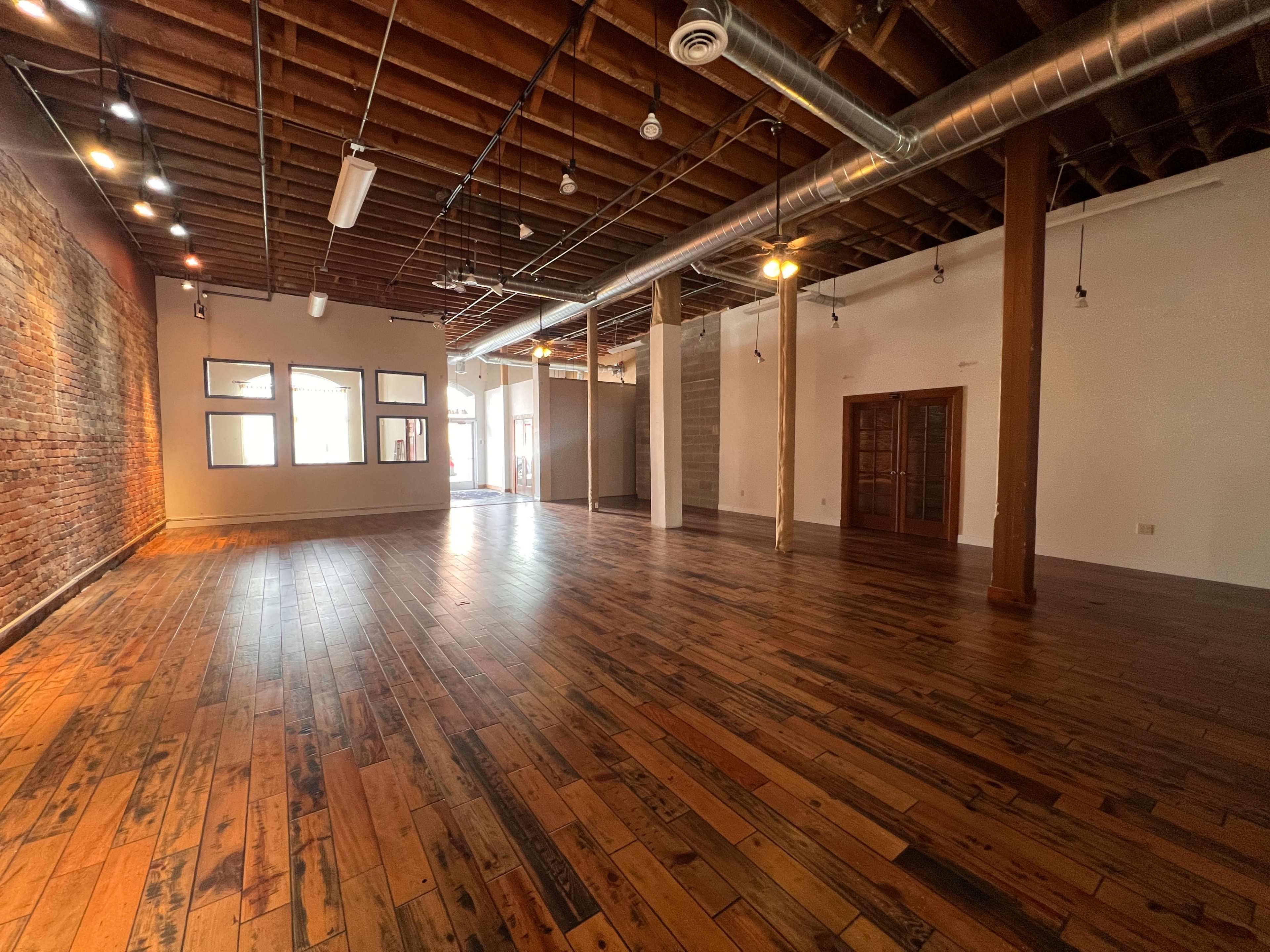 The image shows a spacious, vacant interior room with wooden floors, exposed brick walls, and wooden beams in the ceiling.