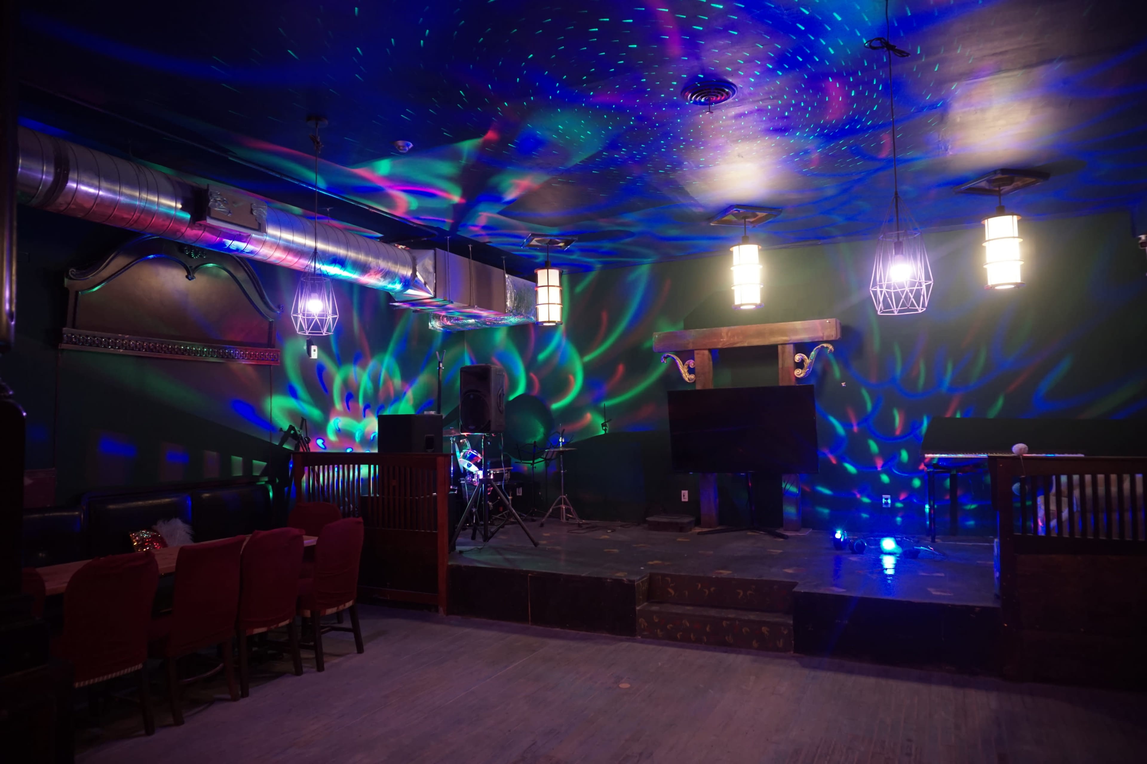 The interior of a dimly lit venue features colorful light patterns on the walls and ceiling, with a stage and musical equipment set up in one corner.