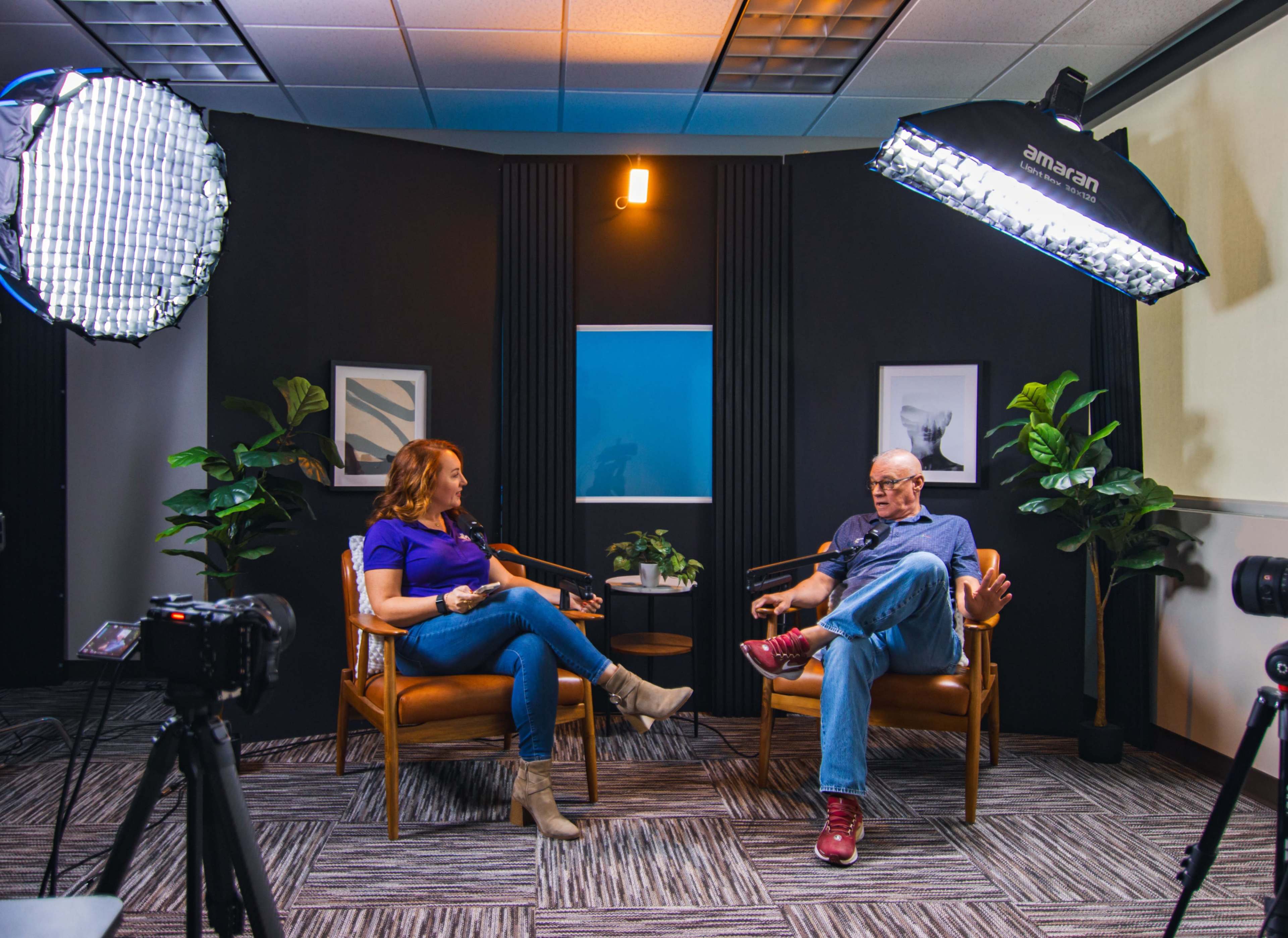 A woman and a man sit in chairs during an interview setup, surrounded by studio lights and green plants.