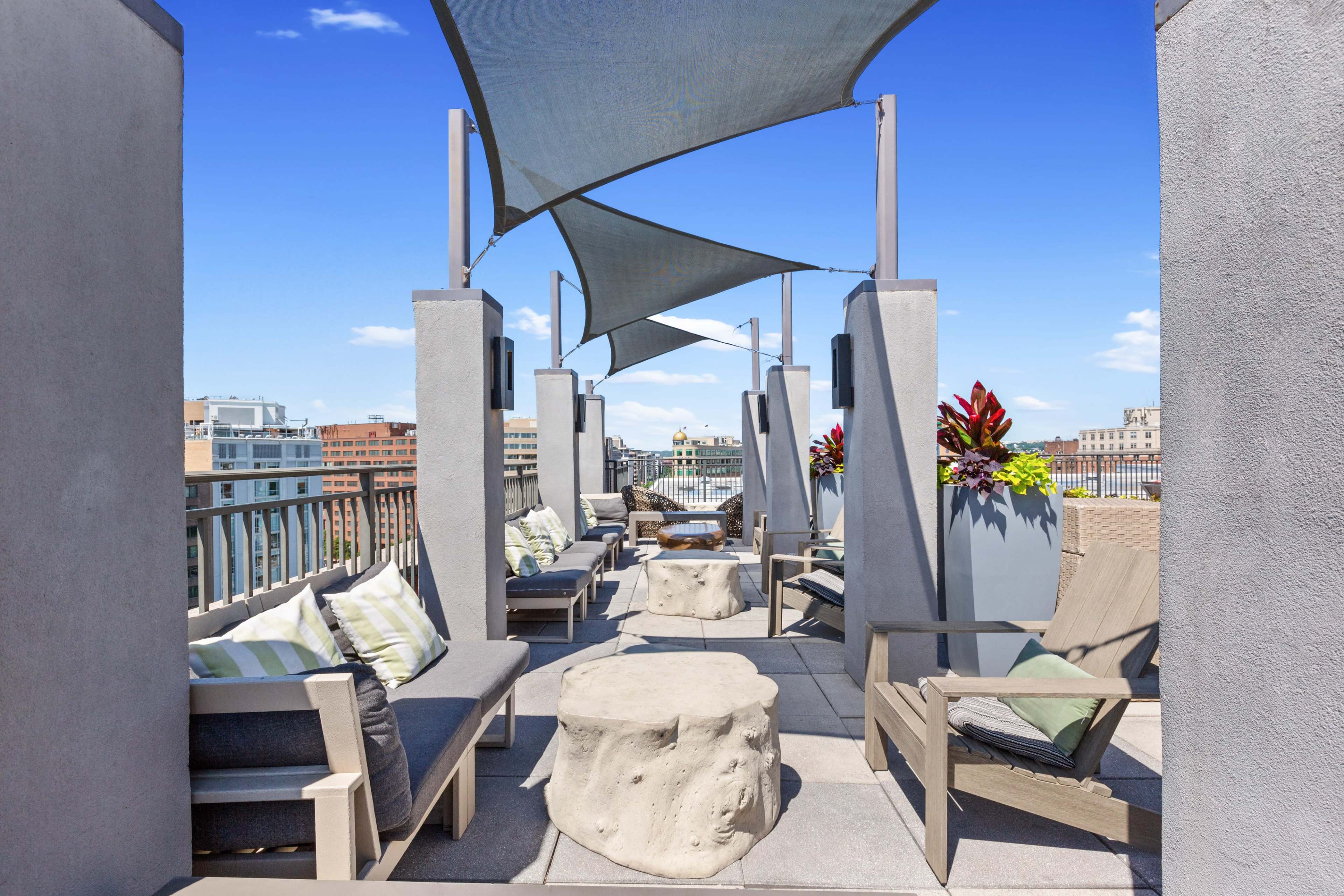 The image shows a rooftop terrace featuring modern seating arrangements, shade sails, and potted plants against a clear blue sky.