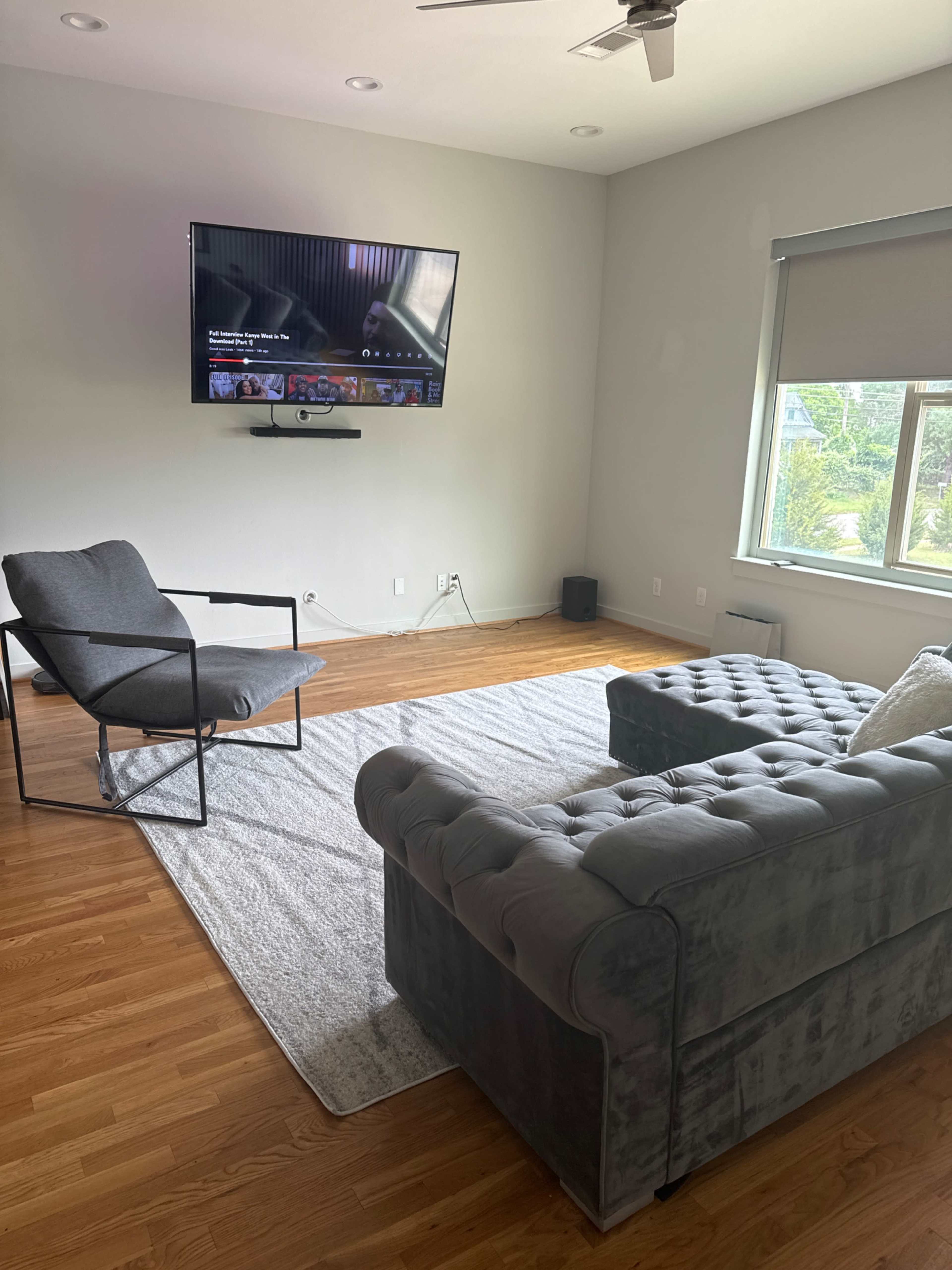 The living room features a gray tufted sofa and a gray chair facing a wall-mounted television over a light-colored rug.