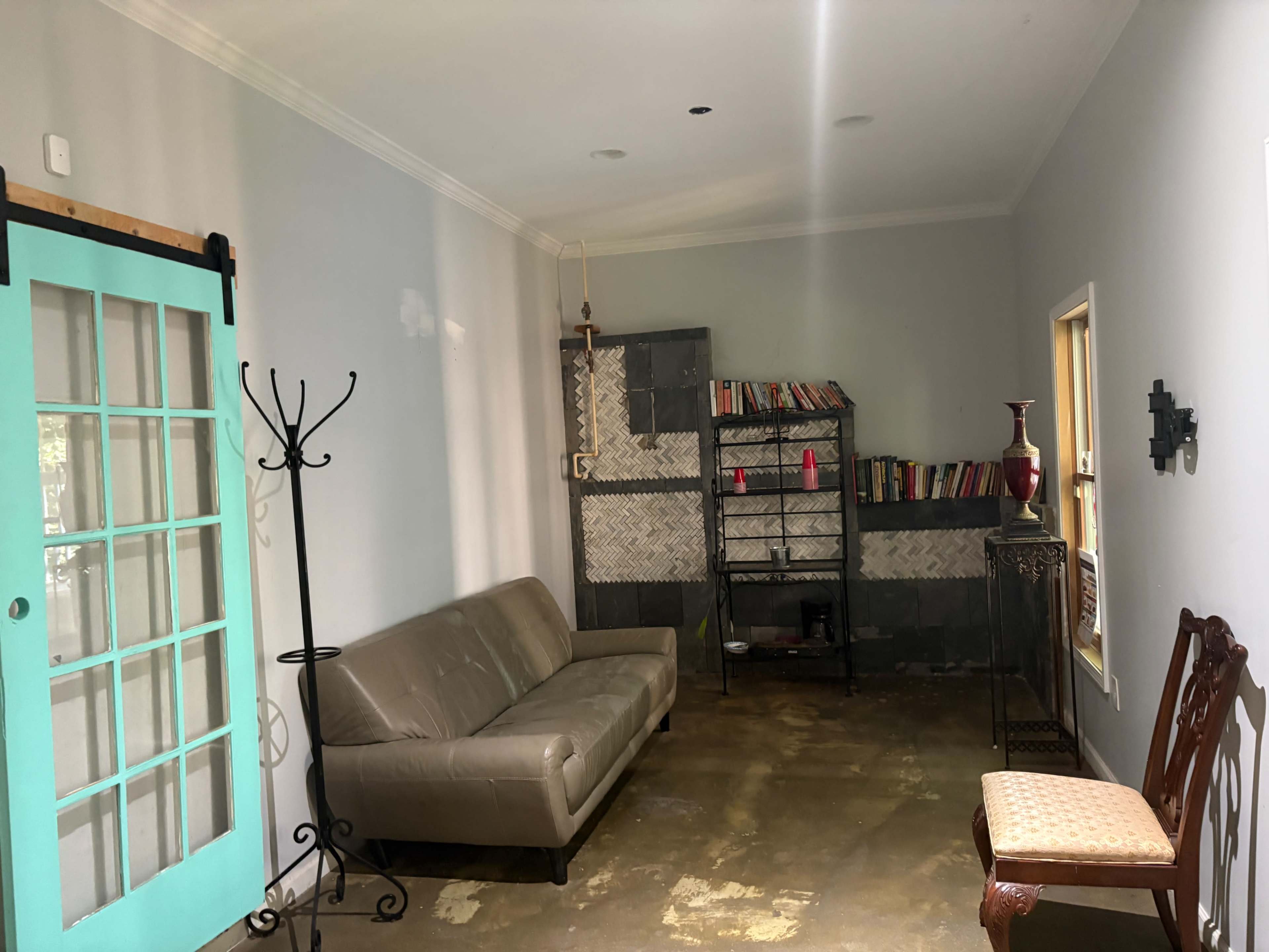 The image shows a minimalist living space featuring a light gray wall, a brown sofa, a metal bookshelf filled with books, and a turquoise door.