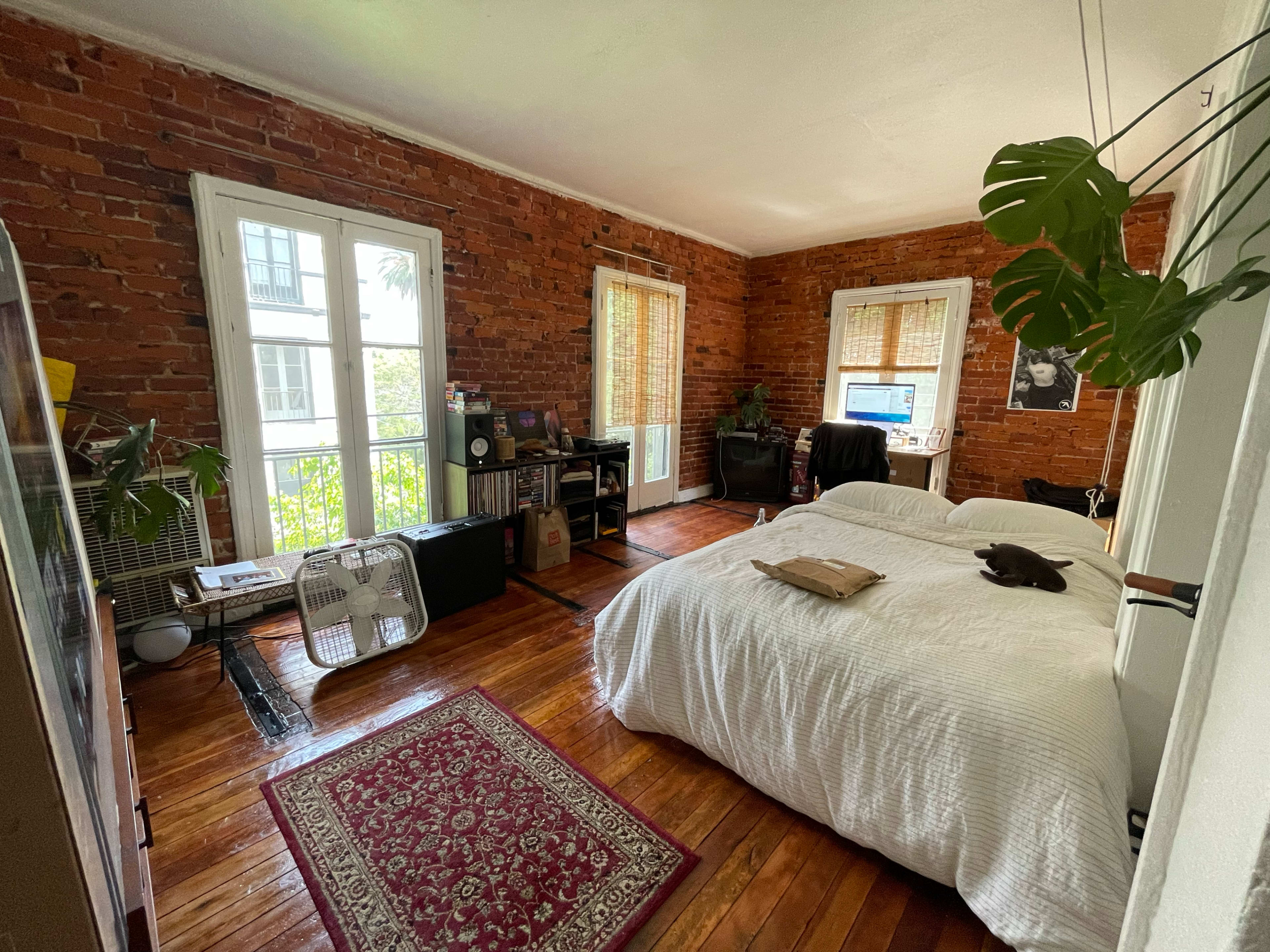 The image shows a bedroom with exposed brick walls, a white bed, wooden flooring, and a desk with a computer near a window.