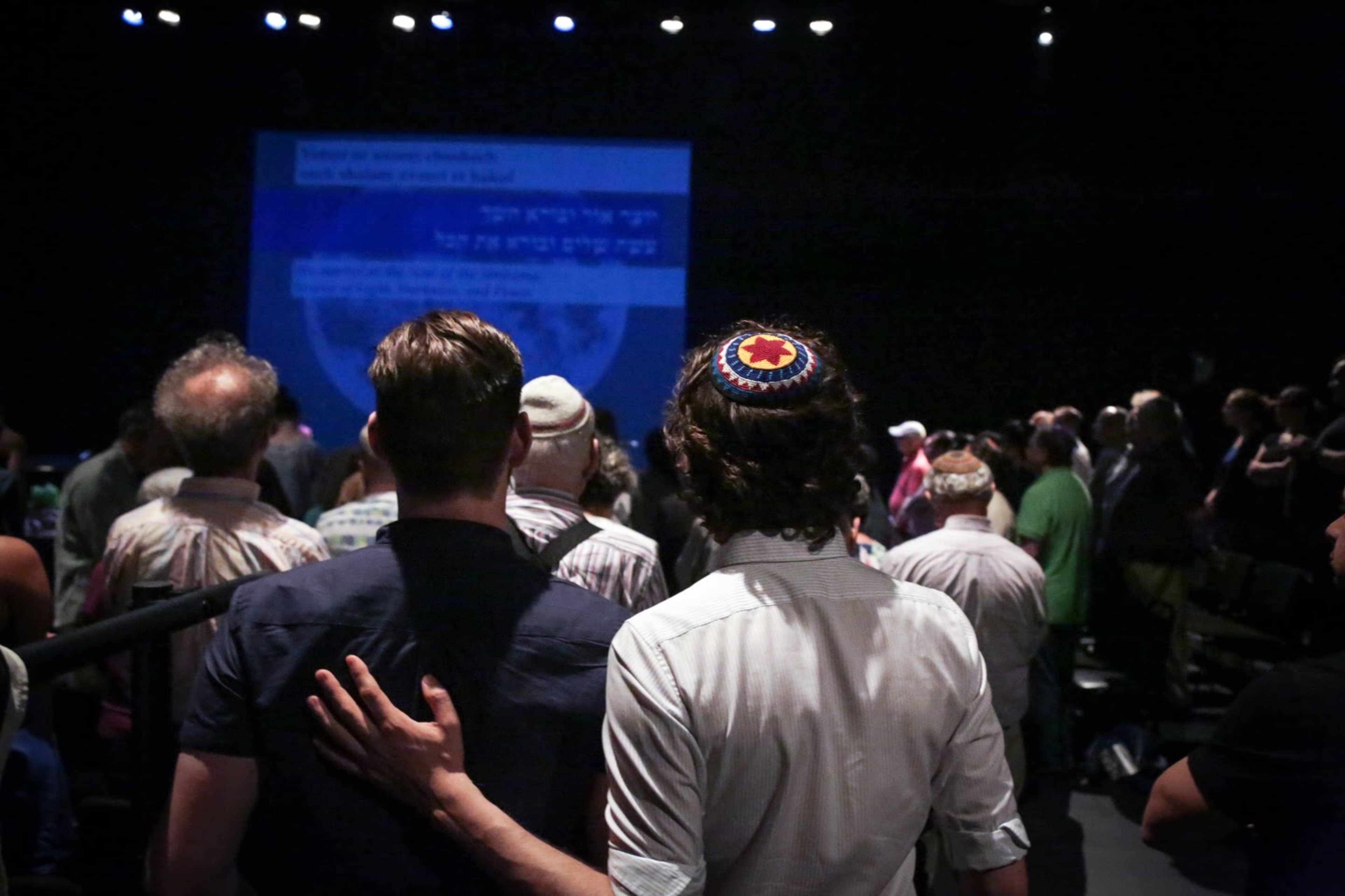 A crowd of individuals stands together in a dimly lit venue, with two men in the foreground facing a presentation screen displaying text in Hebrew.