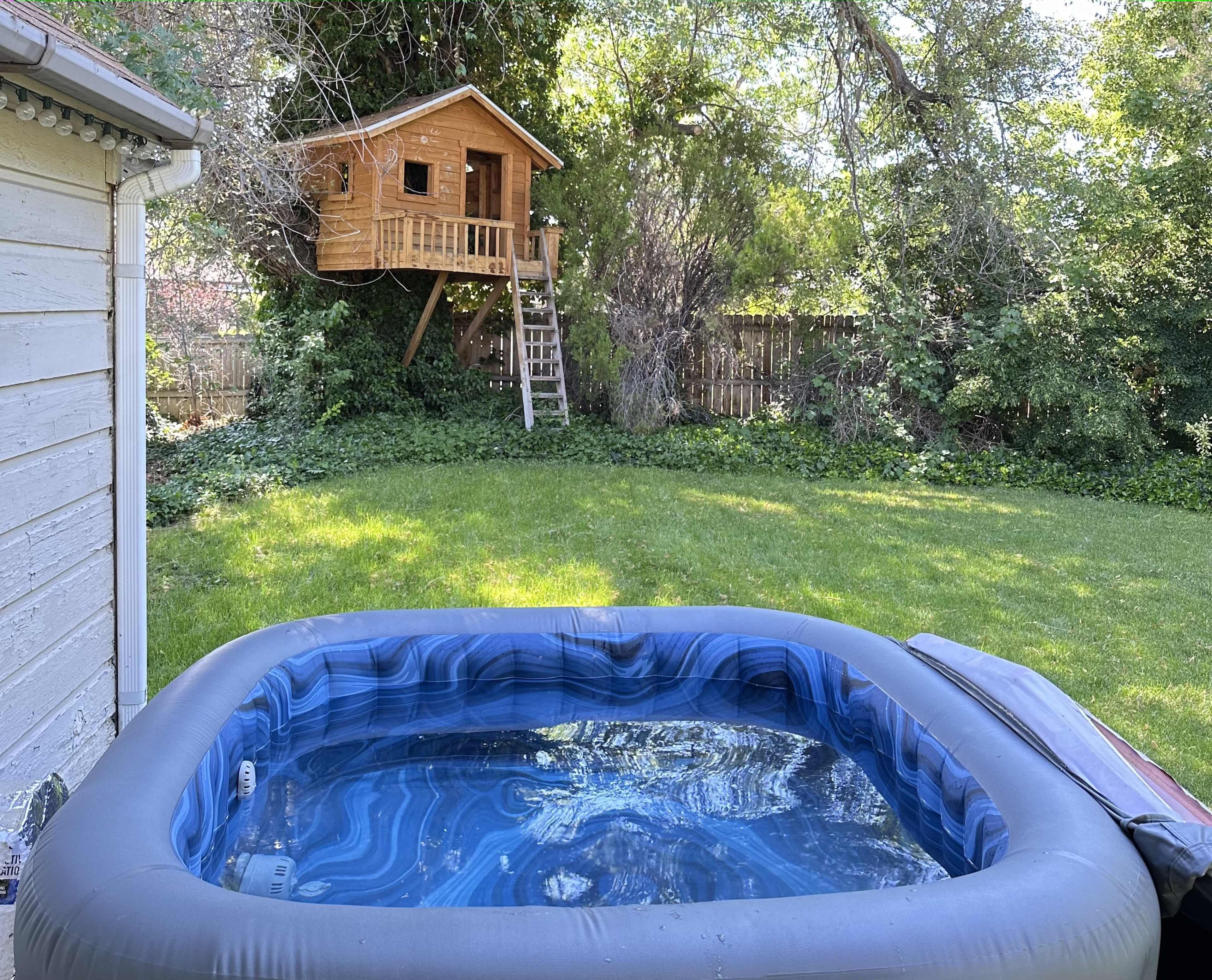 A wooden treehouse is elevated among trees behind a backyard with a hot tub in the foreground.