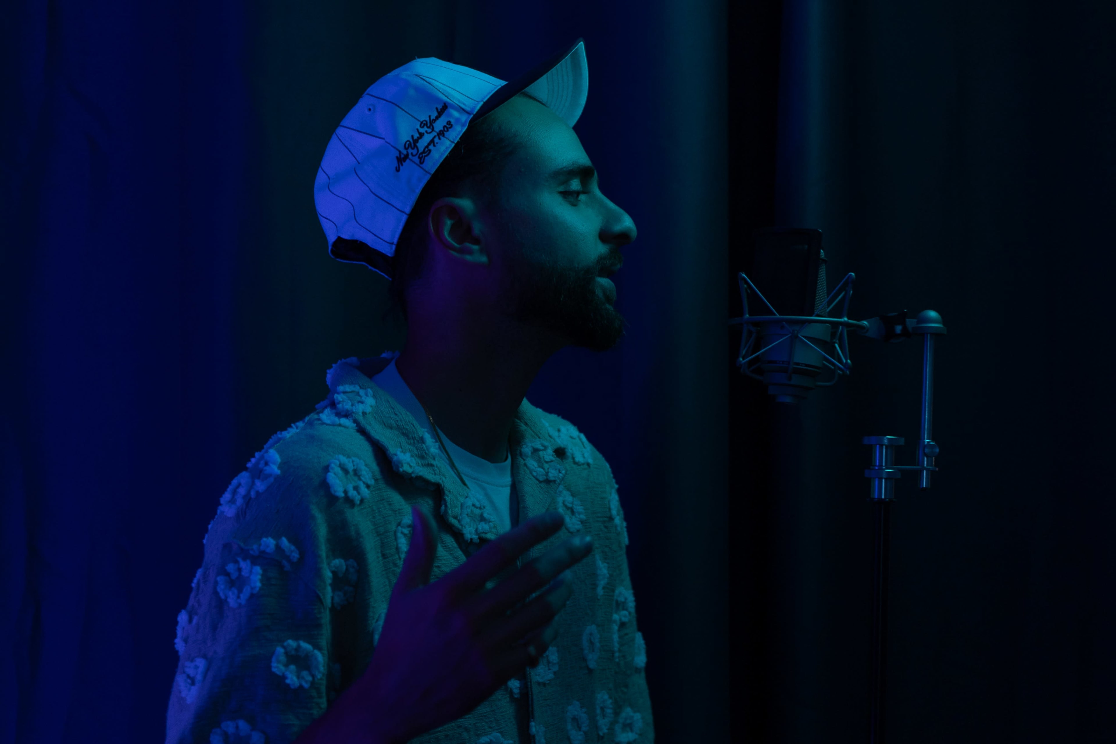 A man wearing a cap stands in profile near a microphone, illuminated by blue lighting.