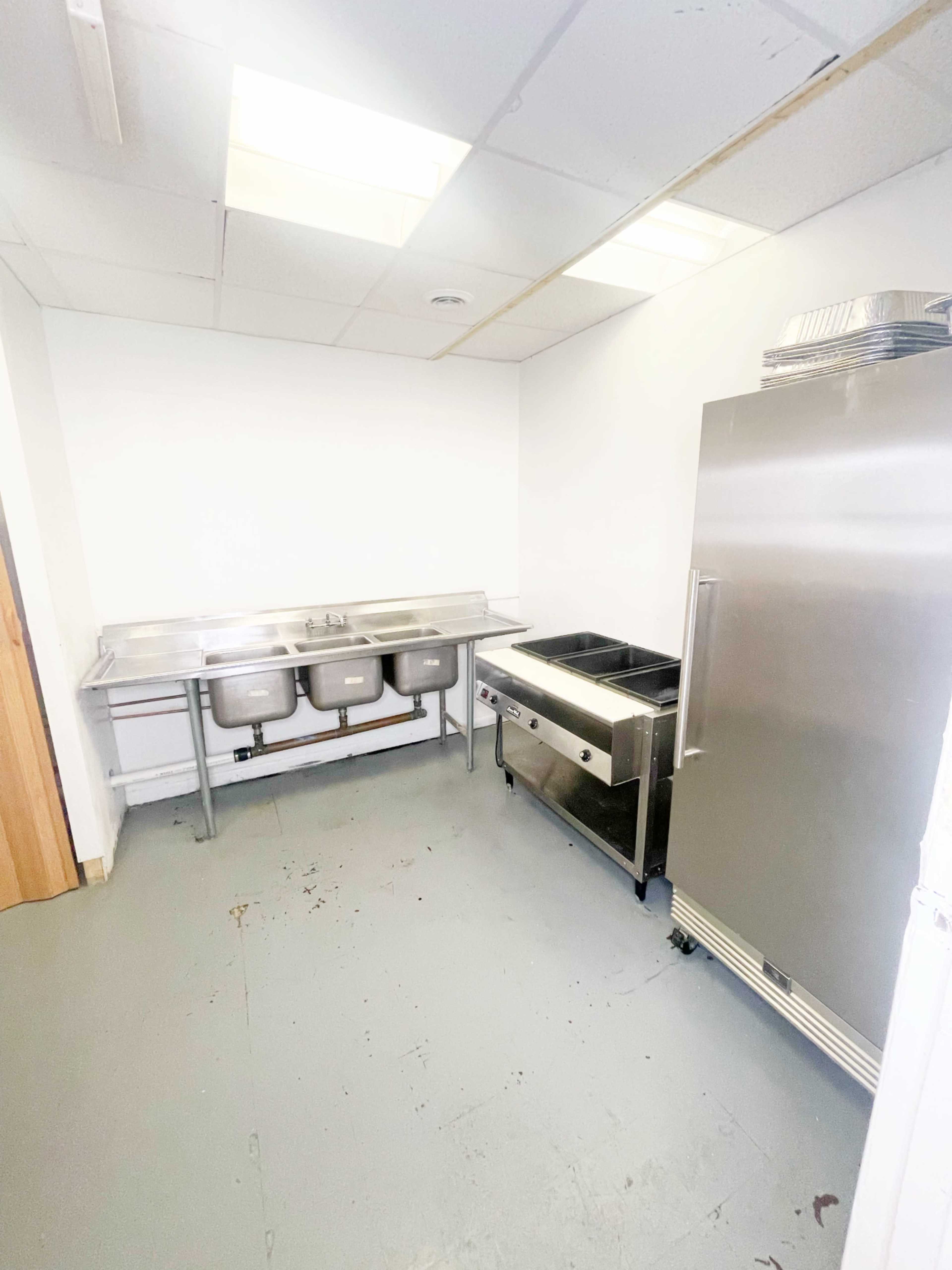A sparse commercial kitchen with a stainless steel sink, a refrigerated unit, and a countertop covered with a serving station.
