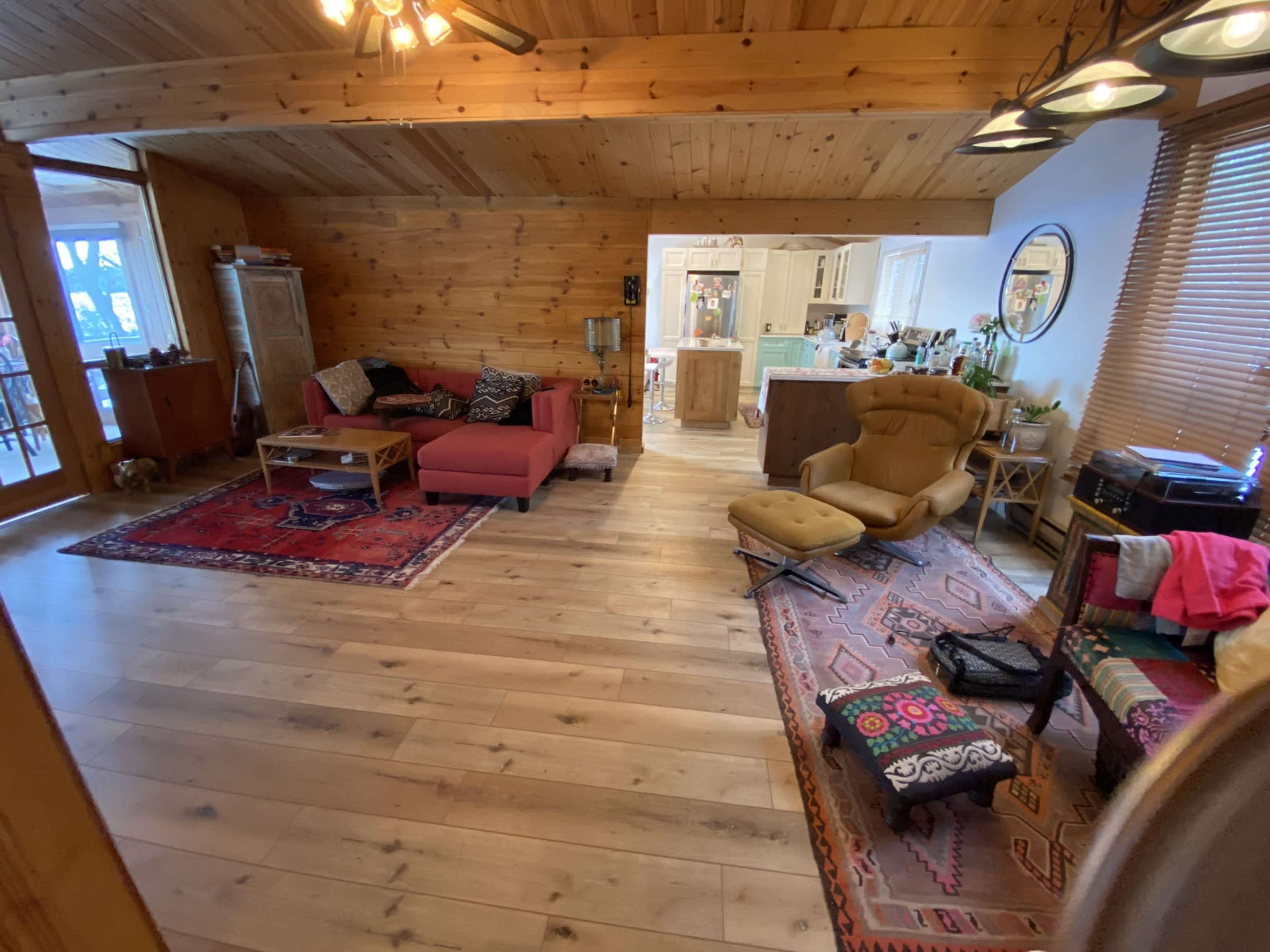 A cozy wooden living room with a red sofa, a vintage chair, and various rugs on a wooden floor, leading to an adjoining kitchen area.