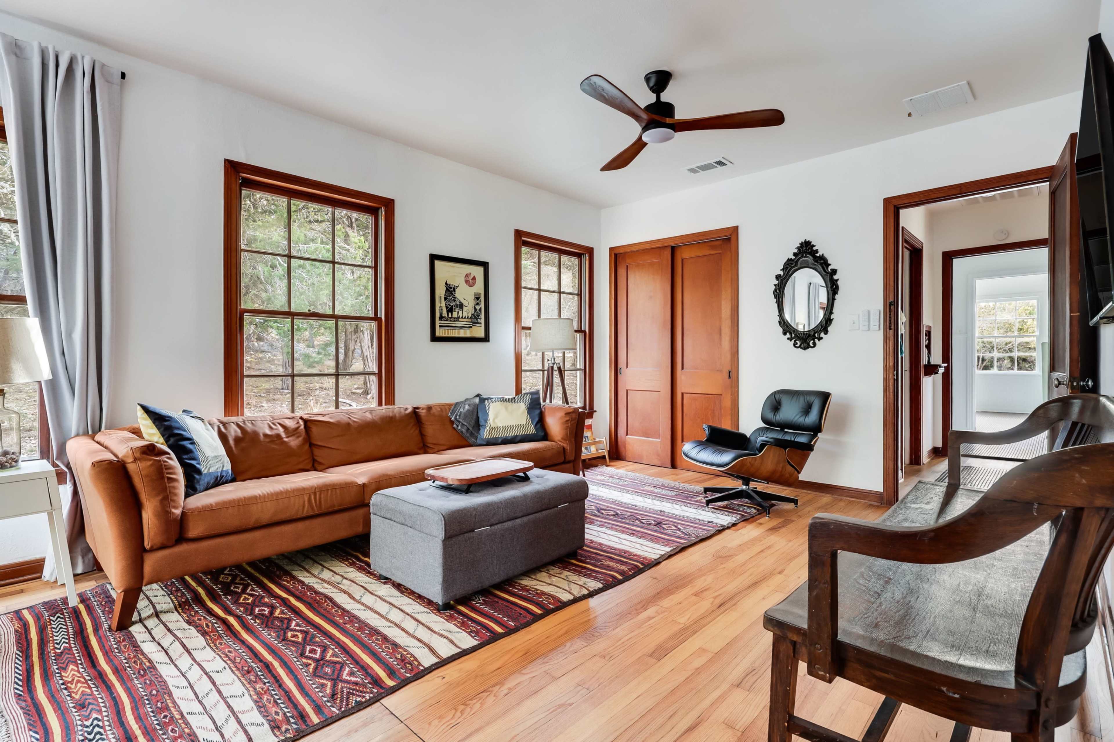 A cozy living room features a brown sofa, a gray ottoman, and large windows, with a patterned rug covering the wooden floor.