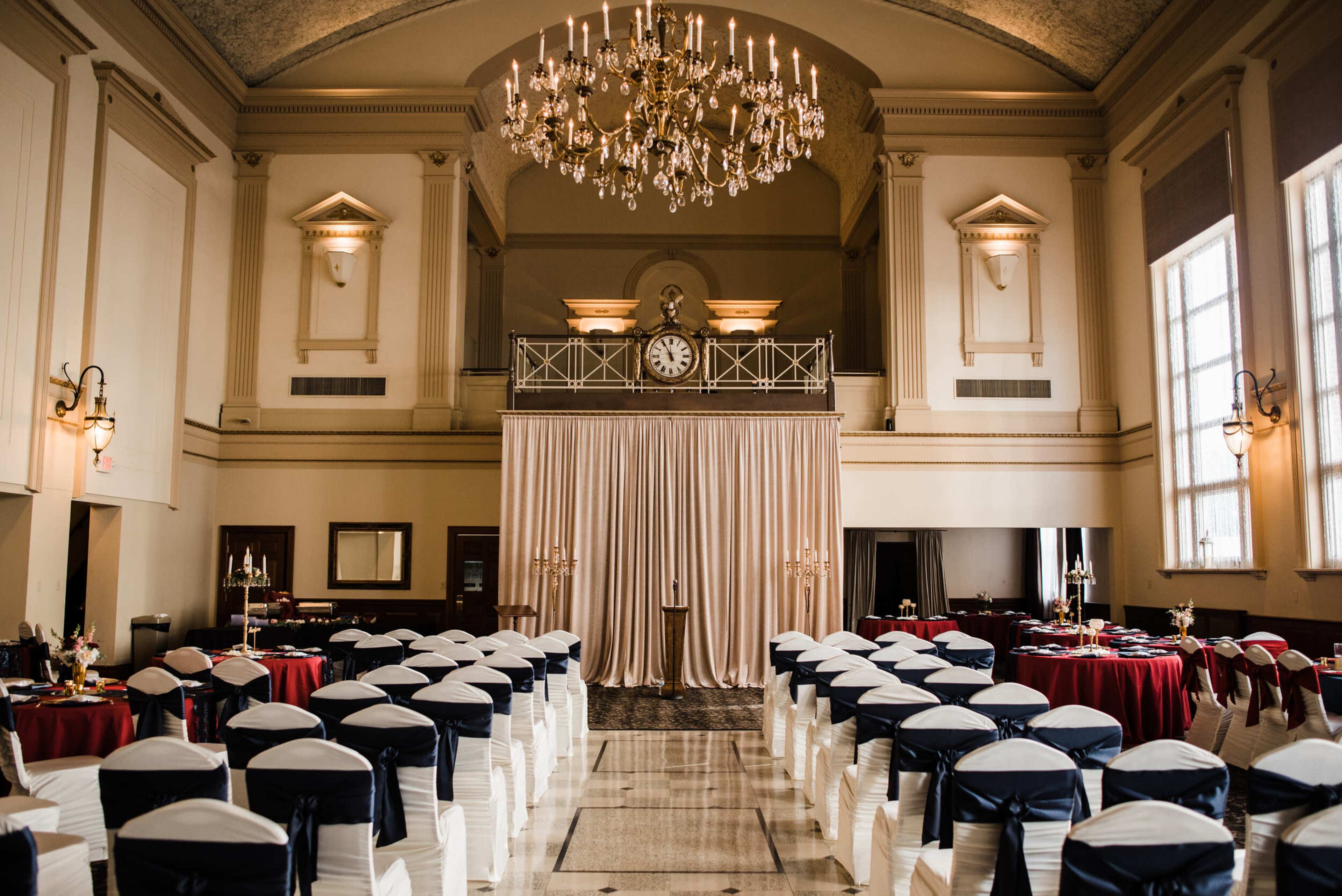 A spacious banquet hall set for an event, featuring rows of chairs draped in white with blue sashes, elegant tables adorned with red tablecloths, and a large chandelier overhead.