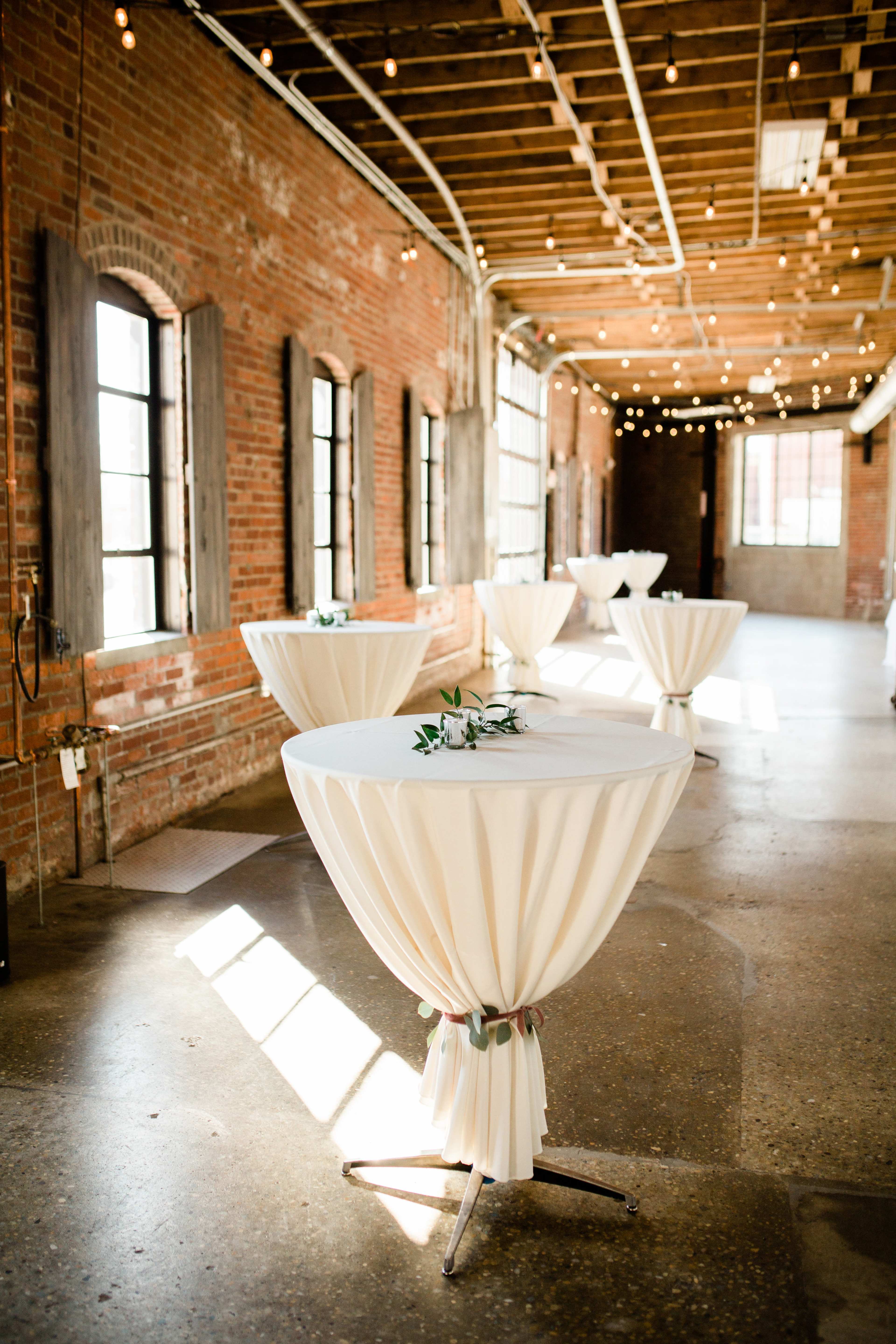 A spacious room with exposed brick walls, adorned with several round tables covered in white tablecloths, illuminated by string lights hanging from the ceiling.