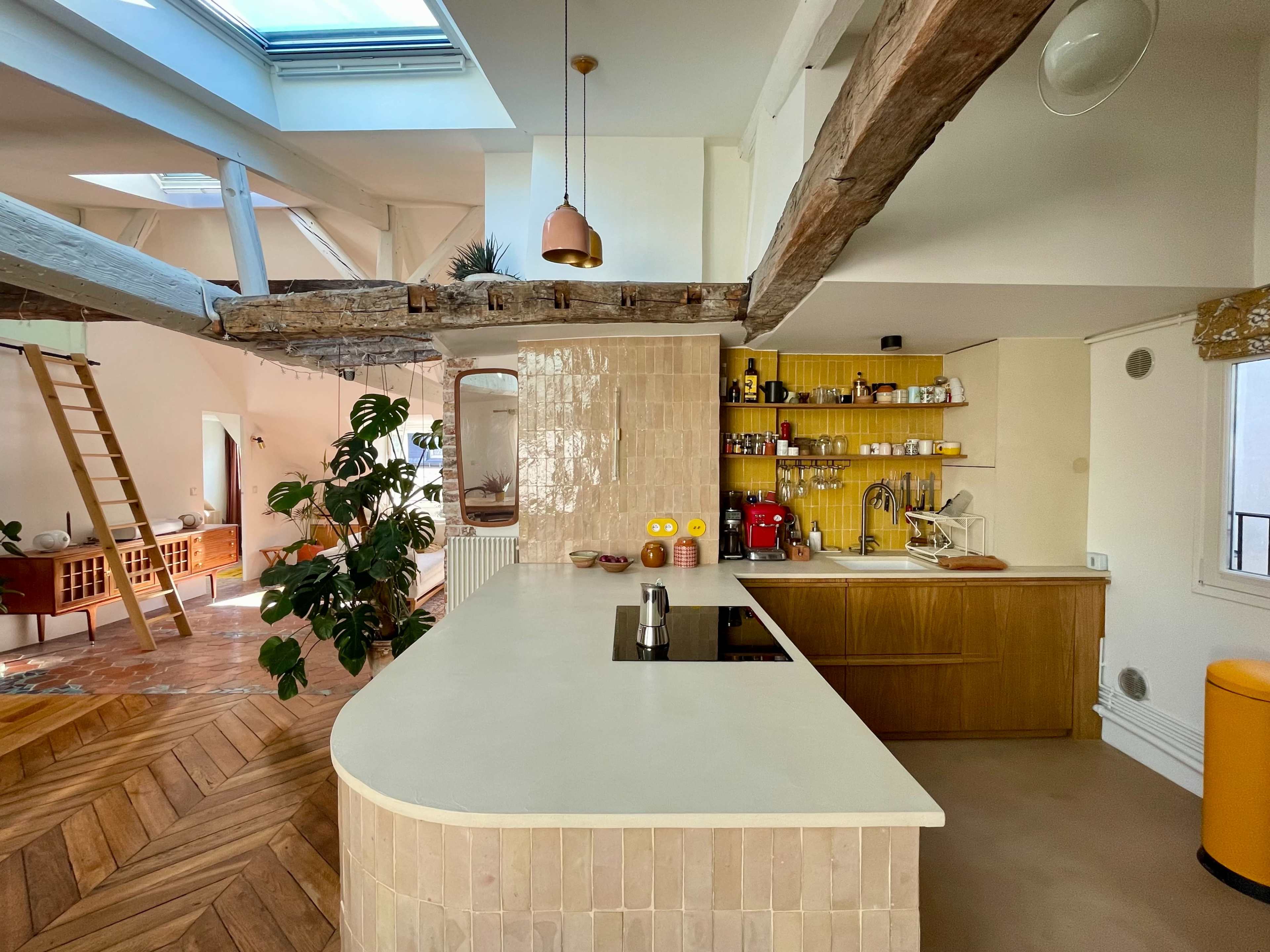 The image shows a modern kitchen with a curved island, wooden beams, yellow walls, and various kitchen appliances and shelves.