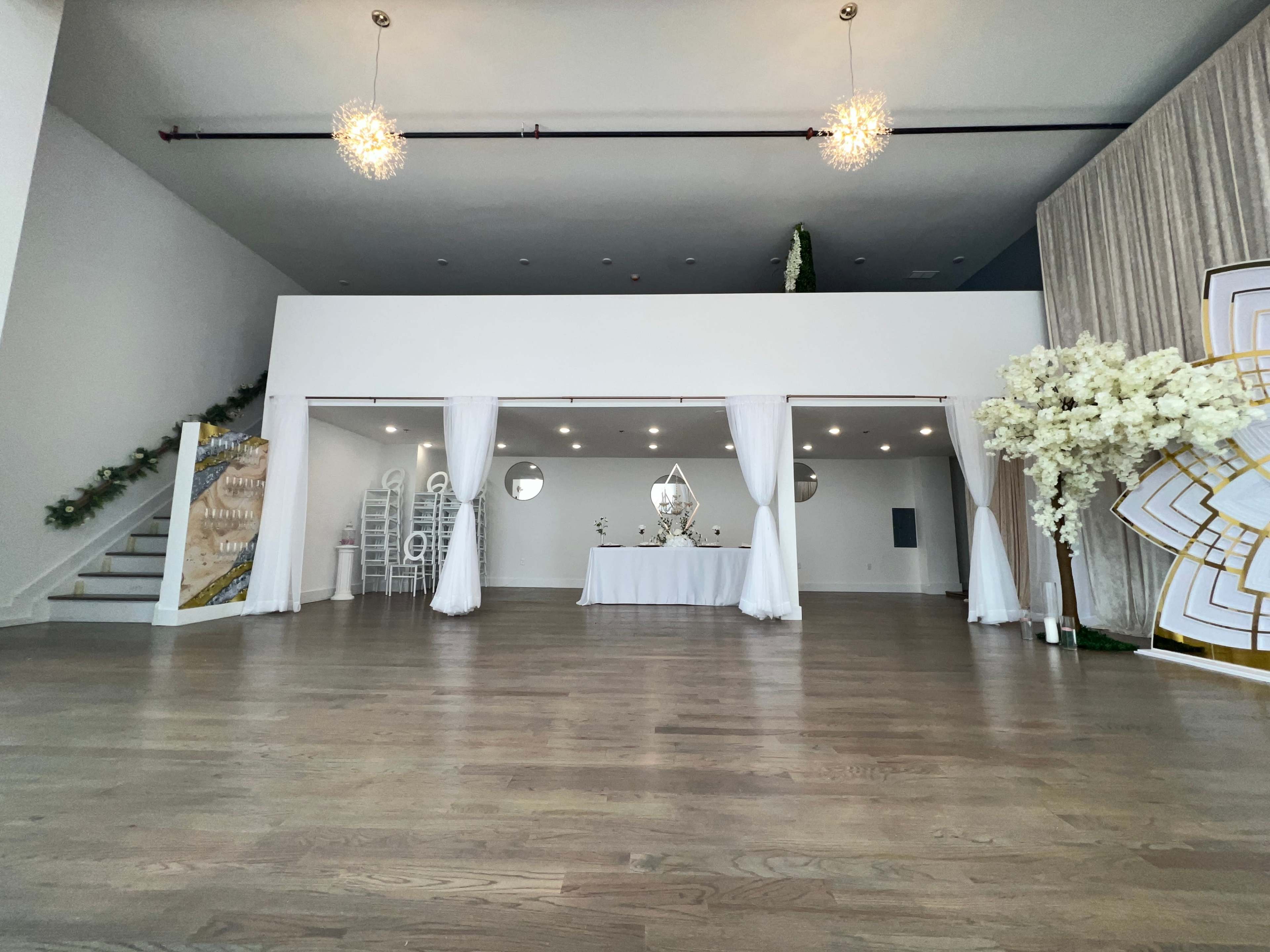 A spacious interior venue with a white draped area, a table set at the center, and elegant decor elements including floral arrangements and lighting fixtures.