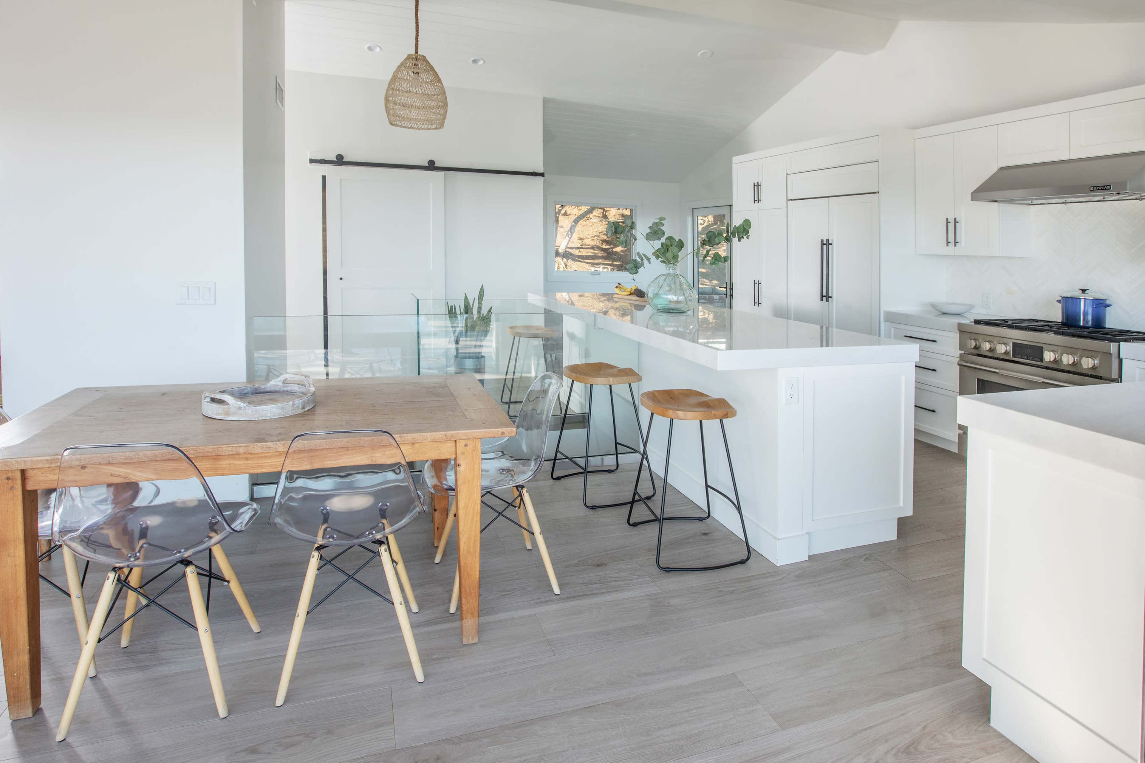 The image shows a modern kitchen with white cabinetry, a central island with bar stools, and a dining table made of wood.