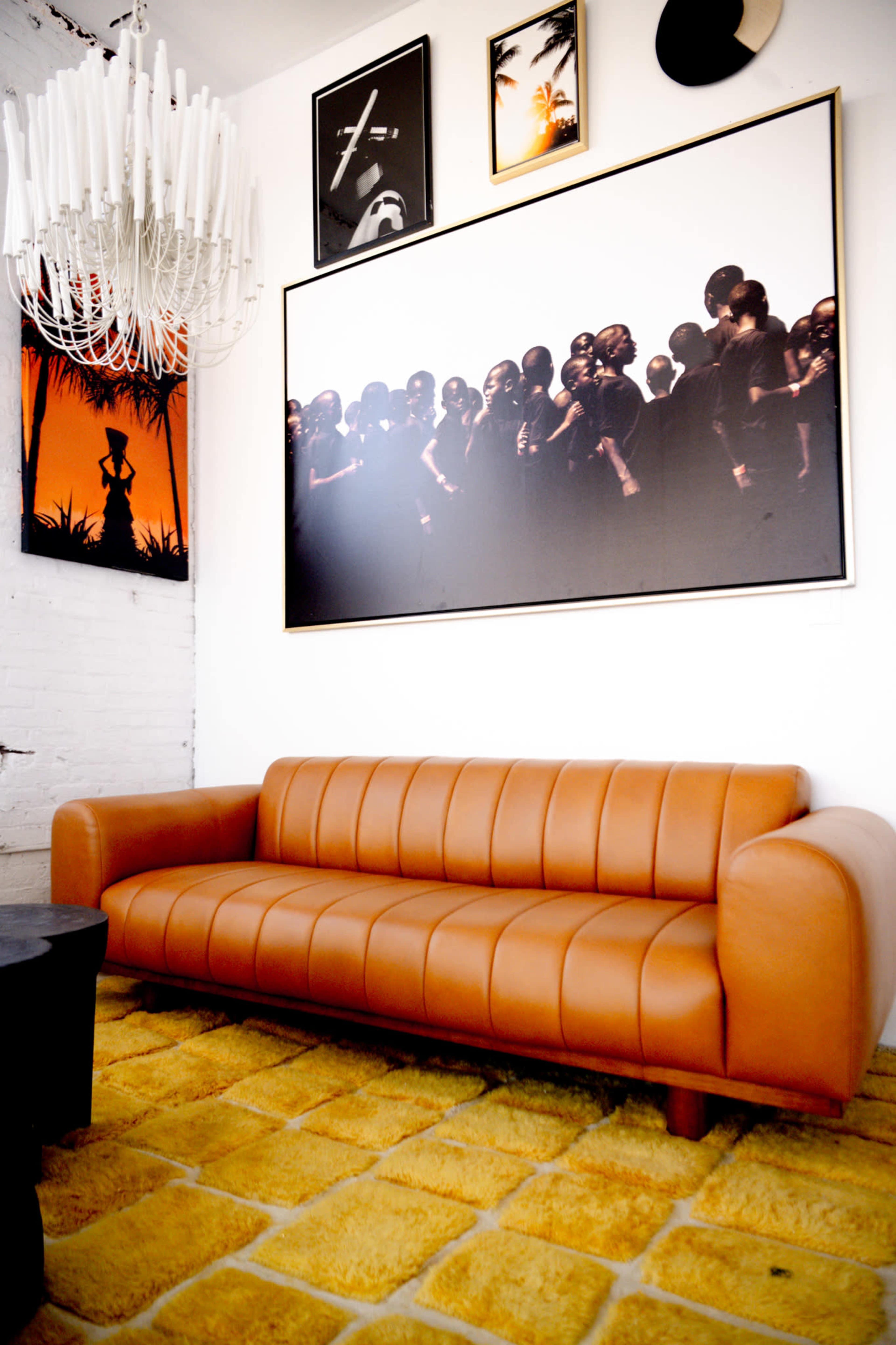 A brown leather sofa is situated on a yellow shag rug below a large framed artwork featuring a group of silhouettes, with additional framed pictures and a chandelier hanging above.