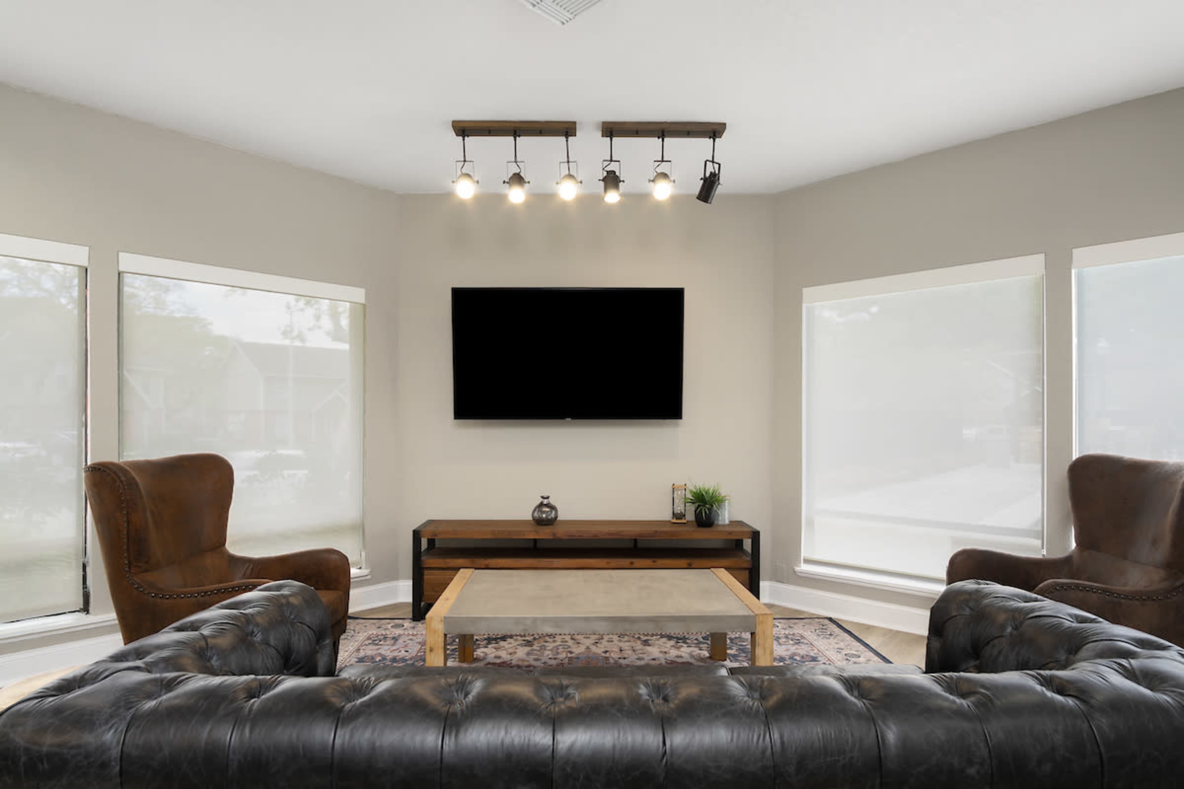 A modern living room features a leather sectional couch, two armchairs, a coffee table, and a wall-mounted television, illuminated by track lighting.