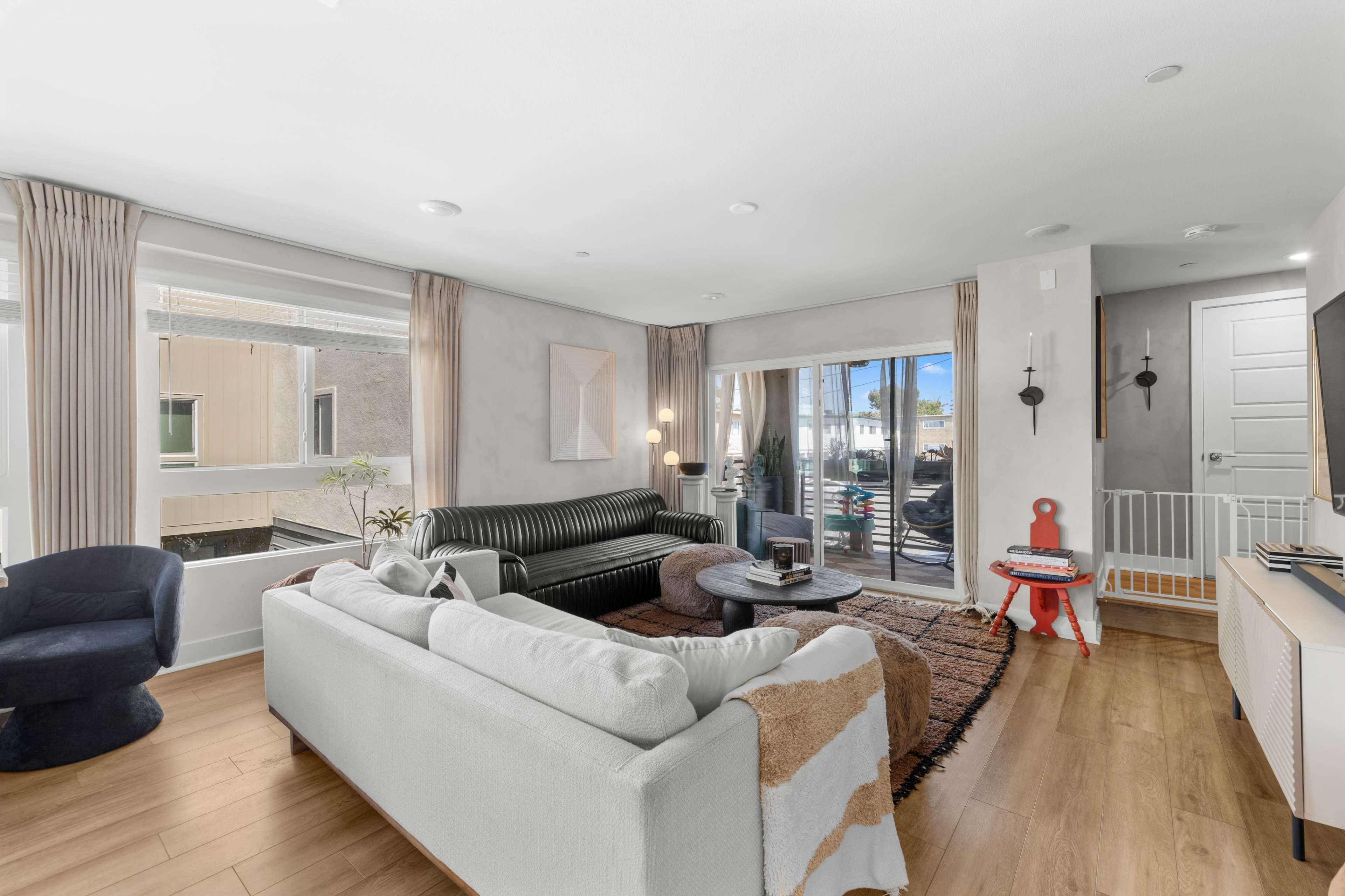 The image shows a modern living room with a black leather sofa, a white sectional couch, large windows, and a coffee table, featuring a mix of light and neutral colors.