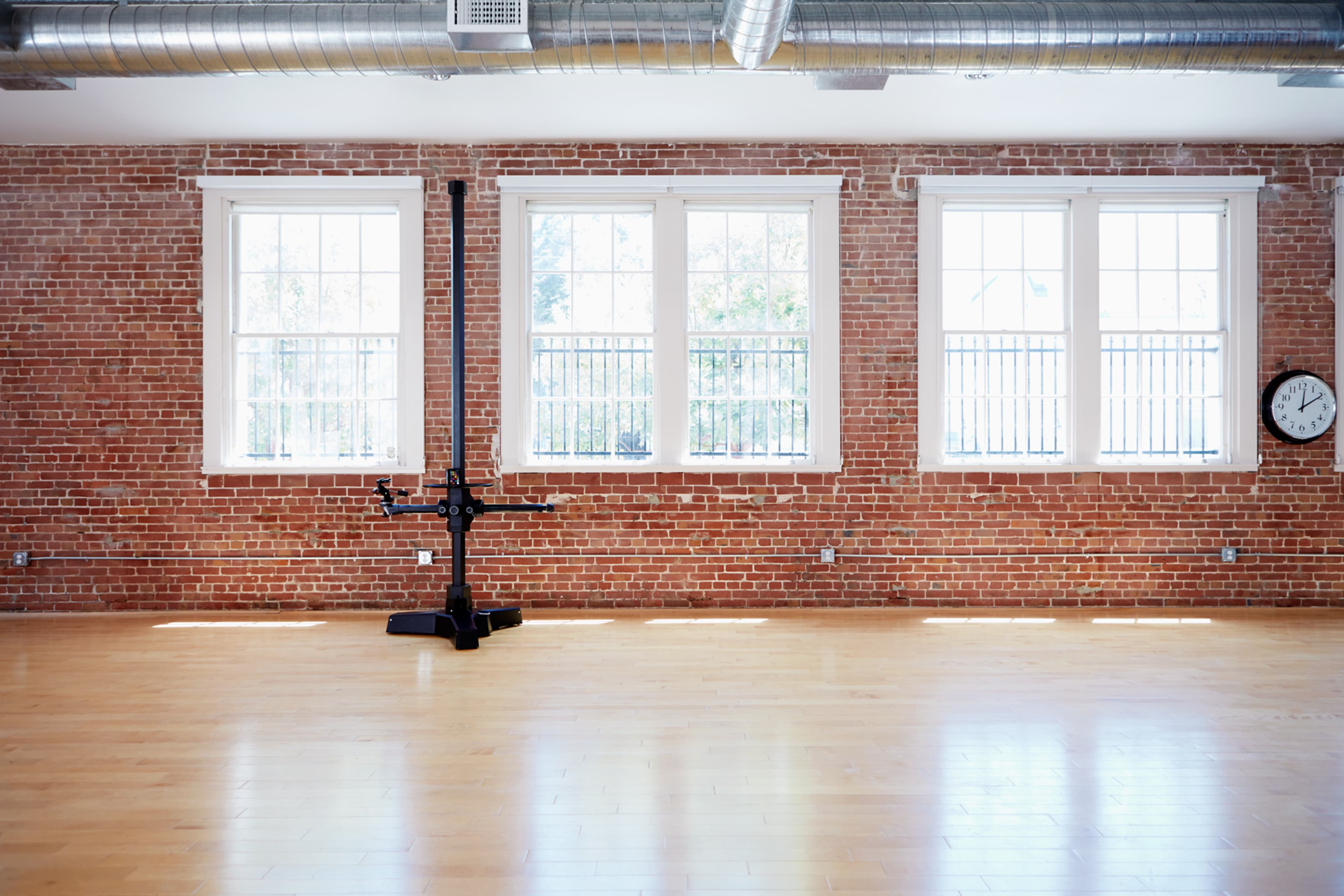 An empty room with hardwood floors, exposed brick walls, large windows, and a clock on the wall.