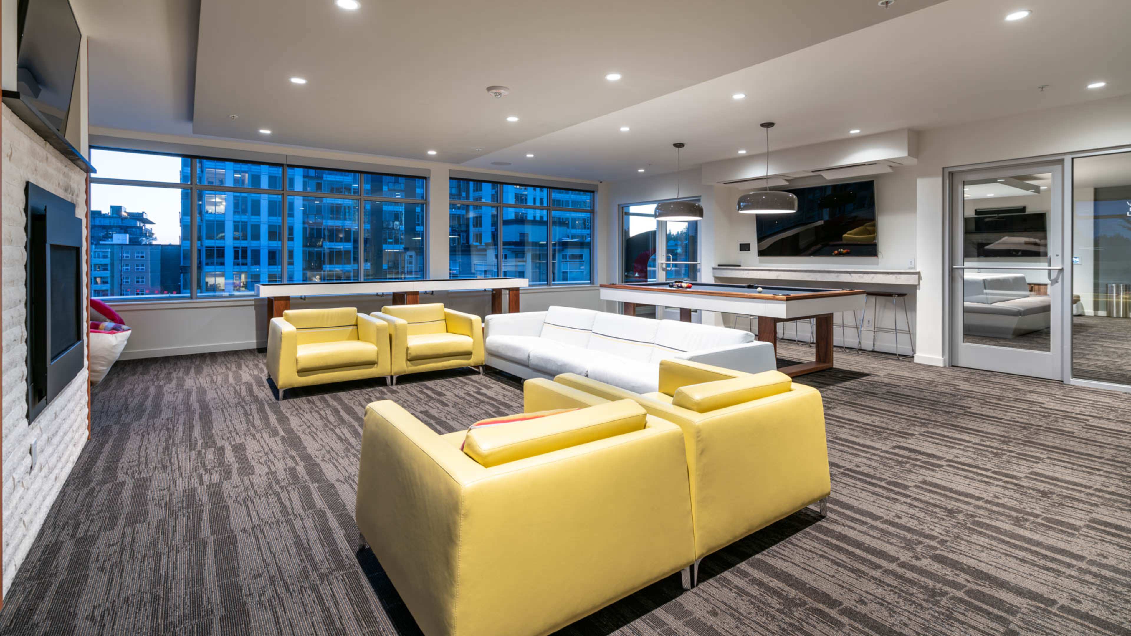 A modern lounge area with yellow and white seating, large windows revealing a cityscape, and a kitchen area in the background.