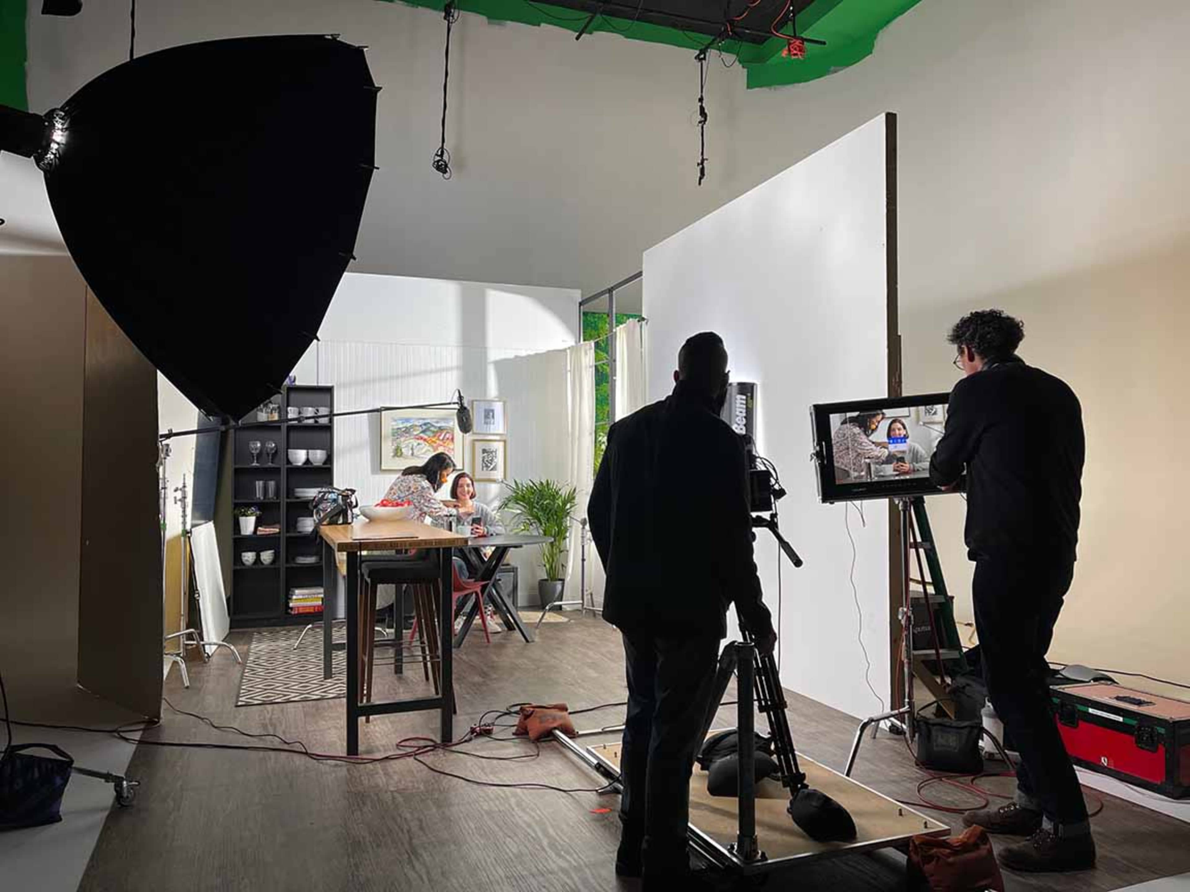 A film set is shown with crew members operating cameras and lighting while a person is seated at a table in the background.