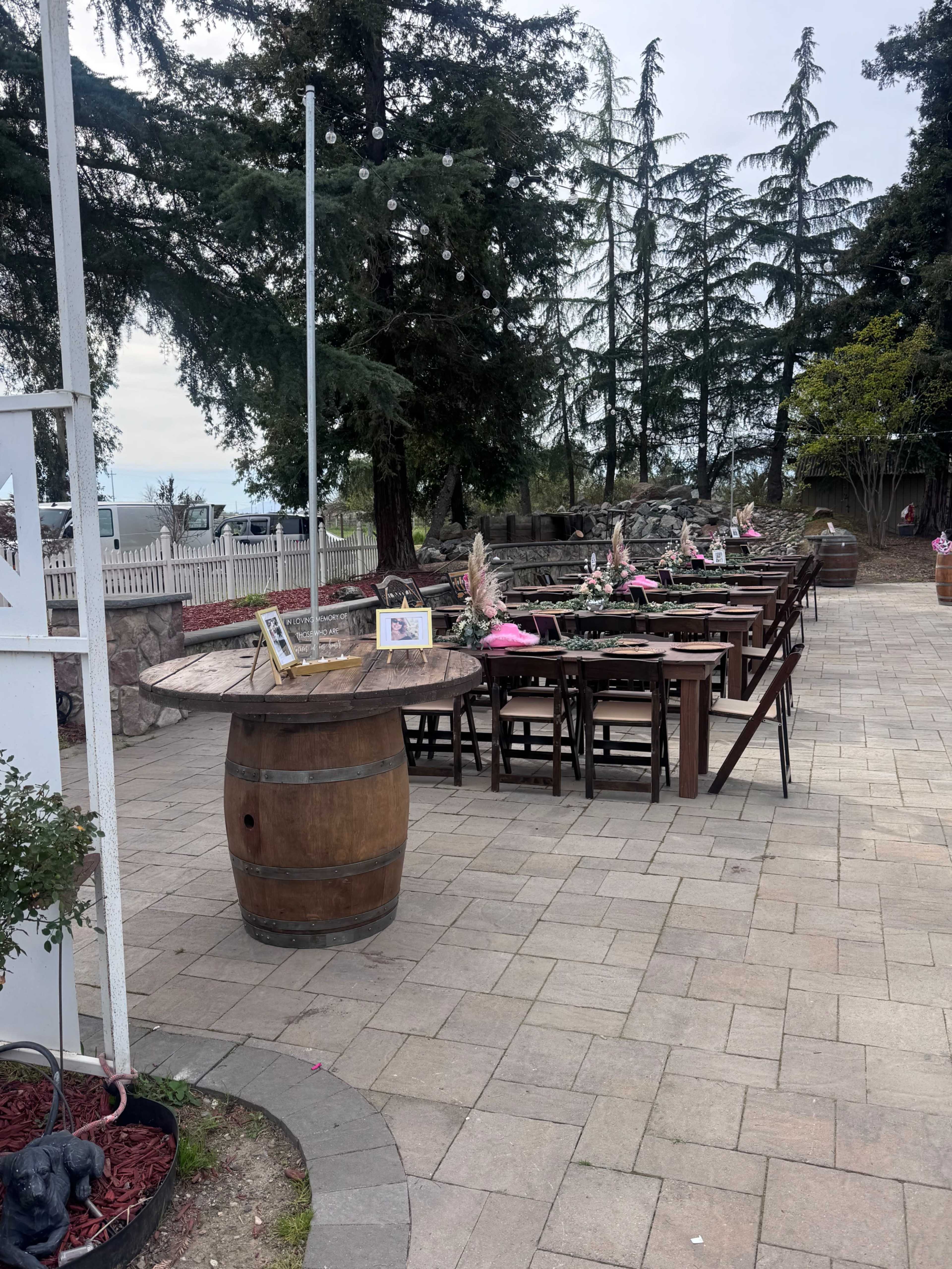 A patio area features wooden tables arranged around a large barrel, with decorative plants and distant trees in the background.