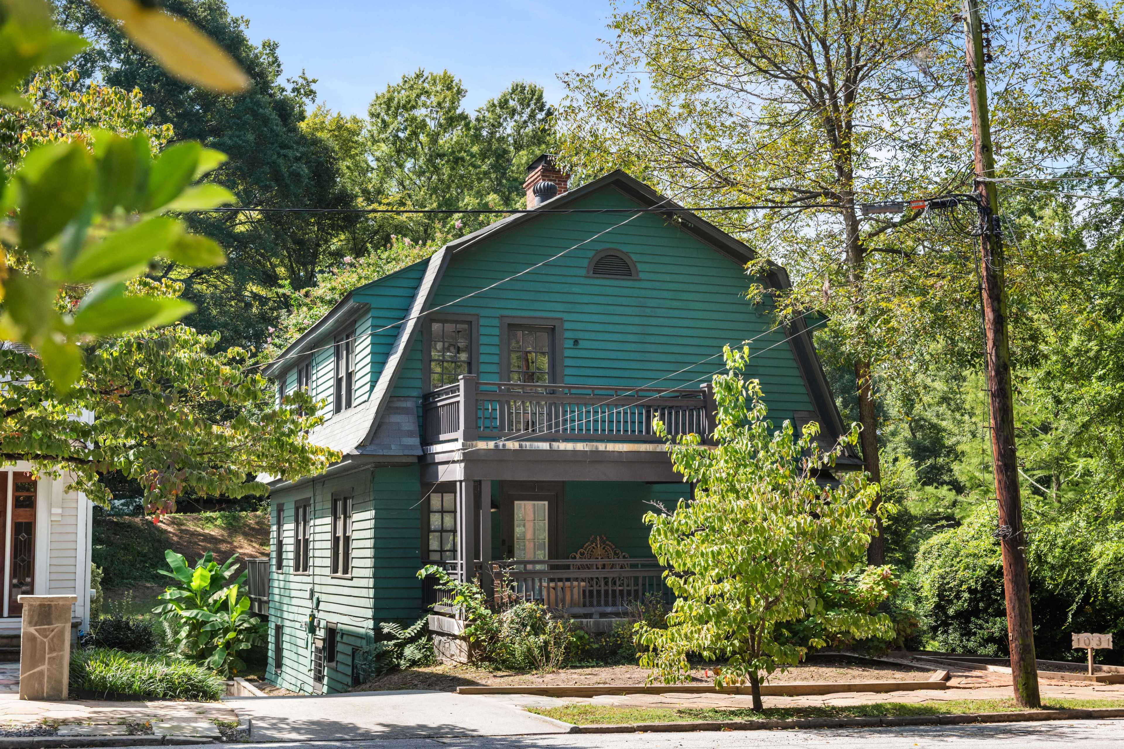 A green, two-story house with a porch and large windows stands among trees in a residential area.