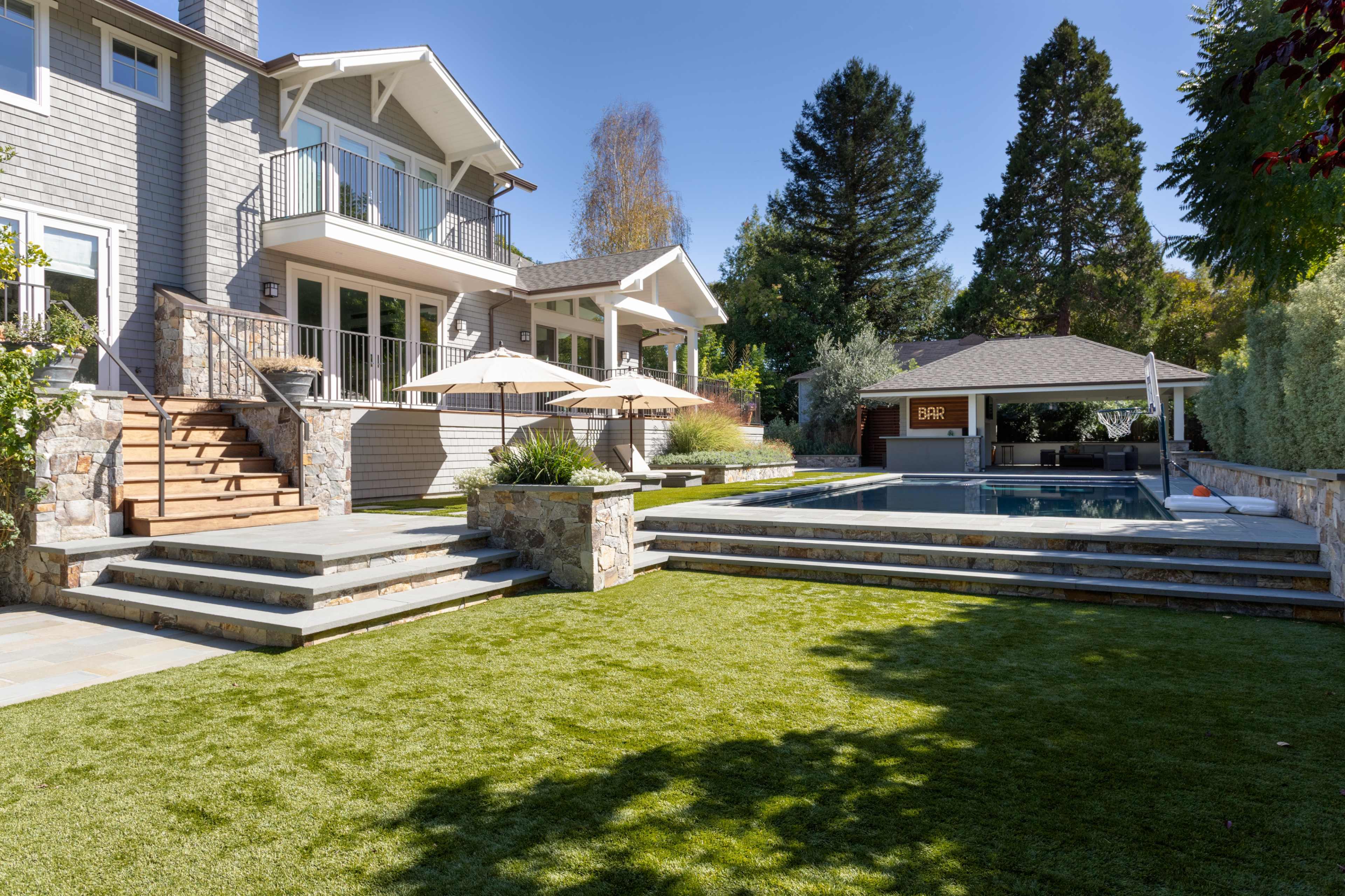A modern home with a landscaped yard, featuring a swimming pool, stone steps, and patio umbrellas.