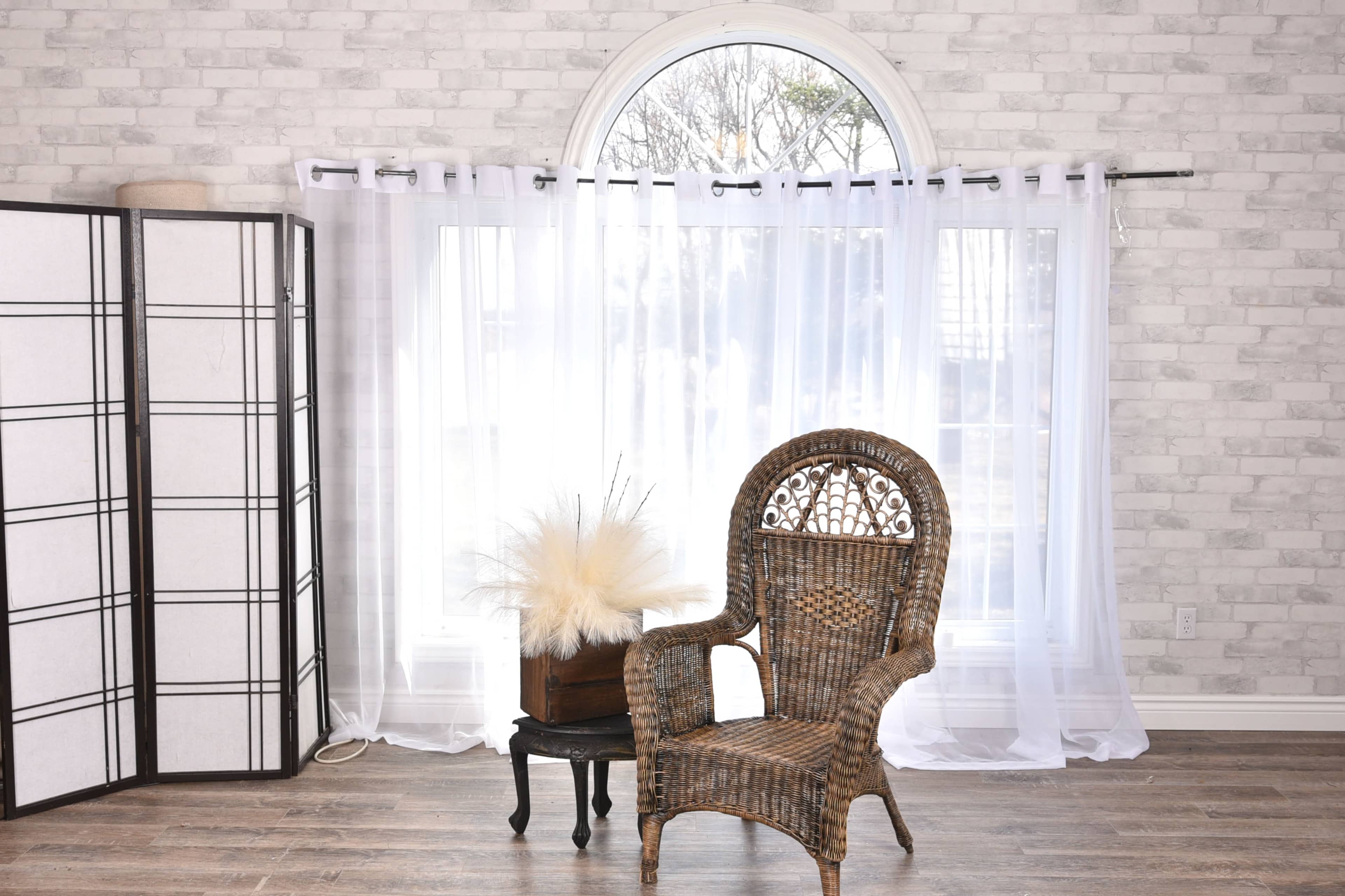 A wicker chair sits beside a small table adorned with a decorative arrangement, set against a large window draped with white curtains in a room with a gray brick wall.