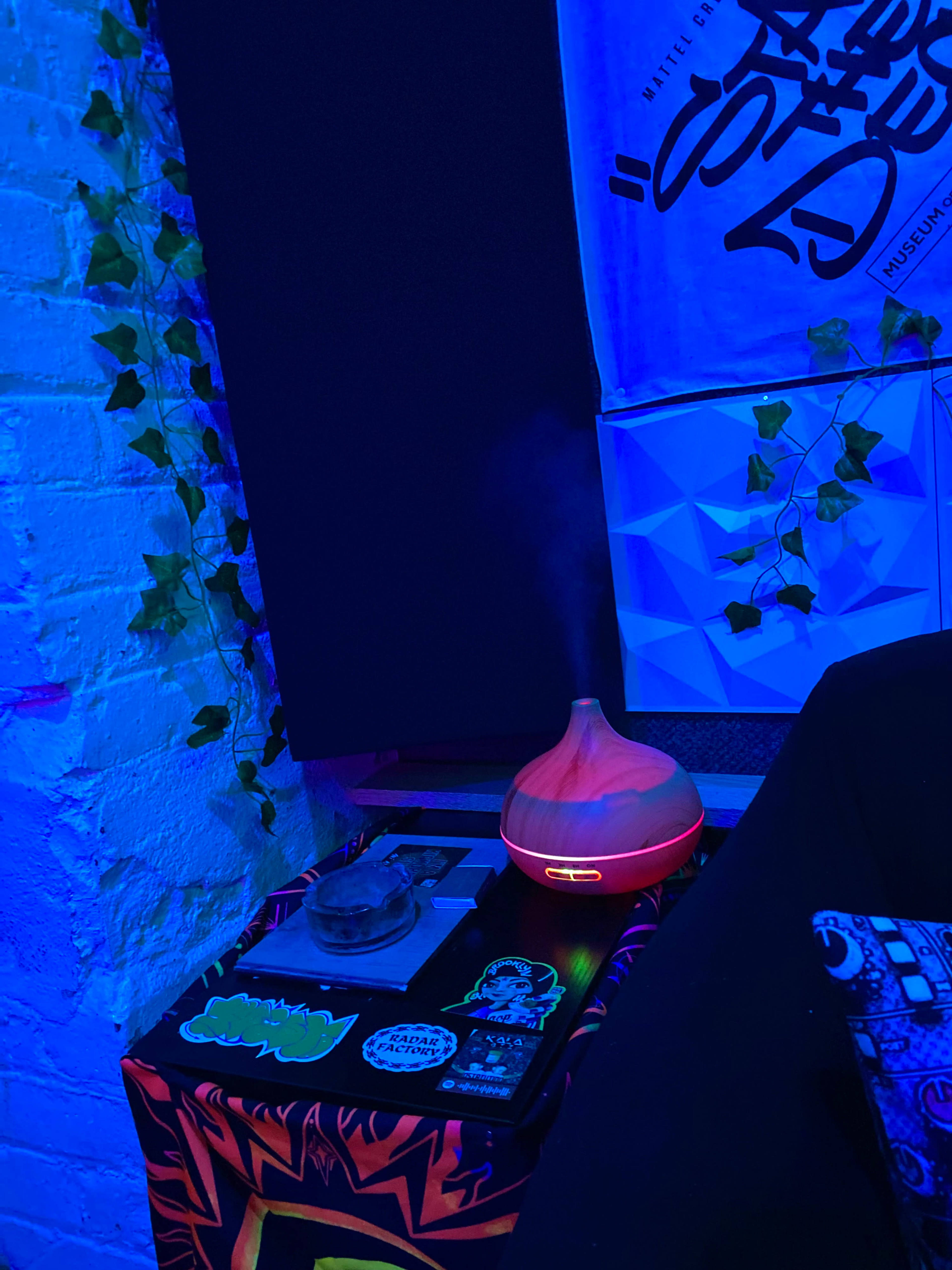 A diffuser emits mist on a table adorned with stickers, next to a plant on a brick wall in a blue-lit room.