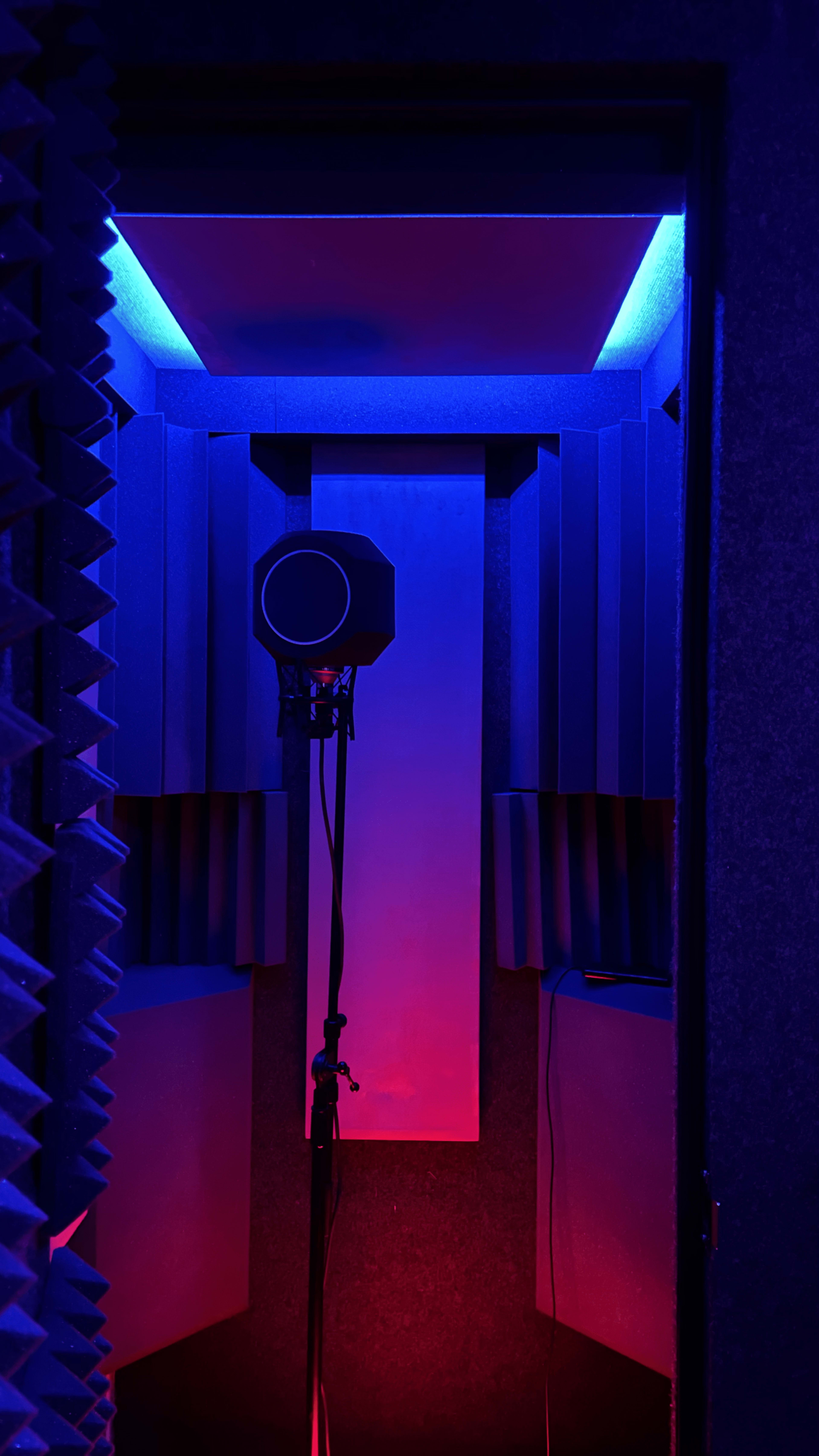 A soundproof recording booth is illuminated with blue and red lighting, featuring a microphone on a stand.