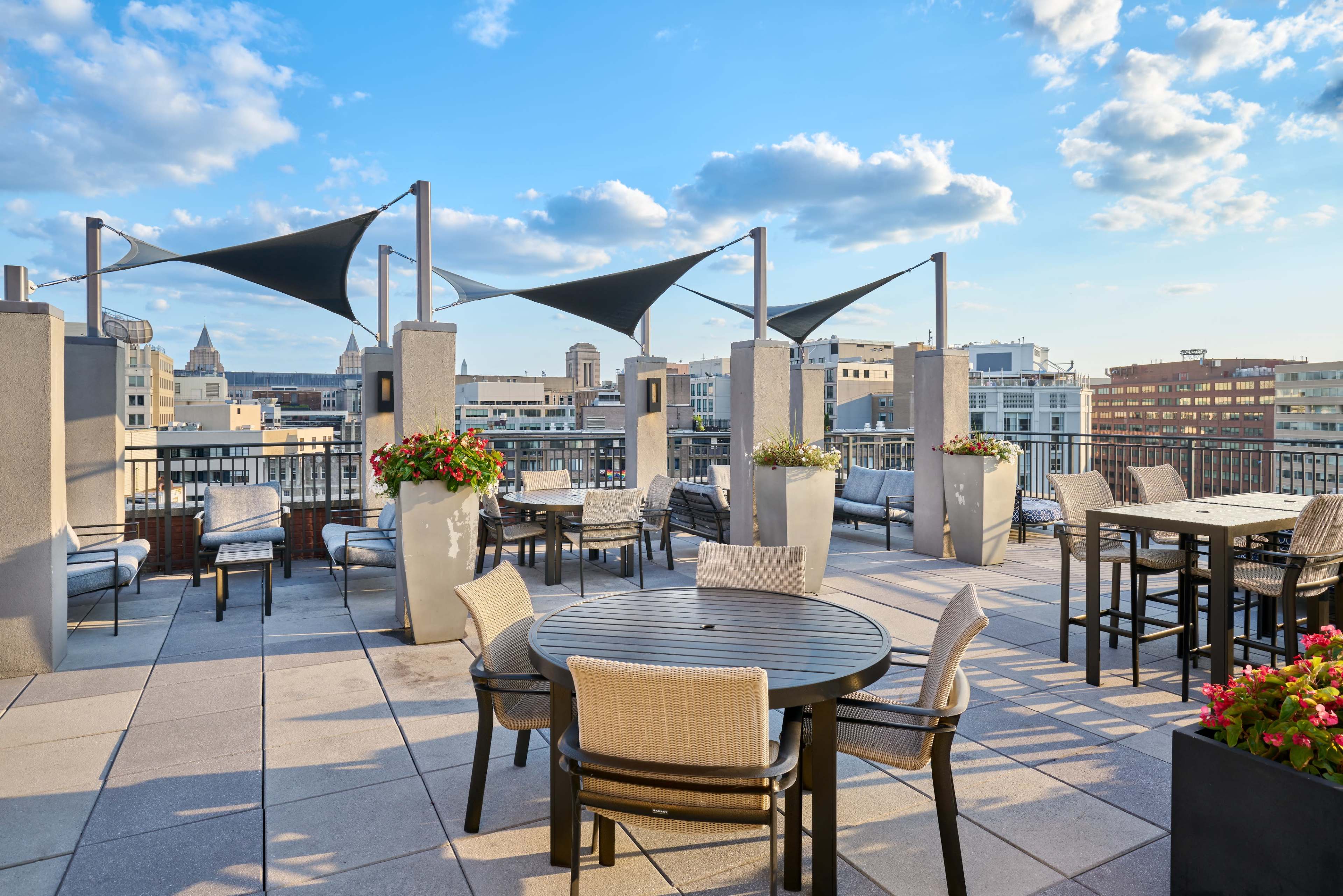 The rooftop patio features several tables and chairs, decorative planters, and shaded areas with a view of the city skyline.