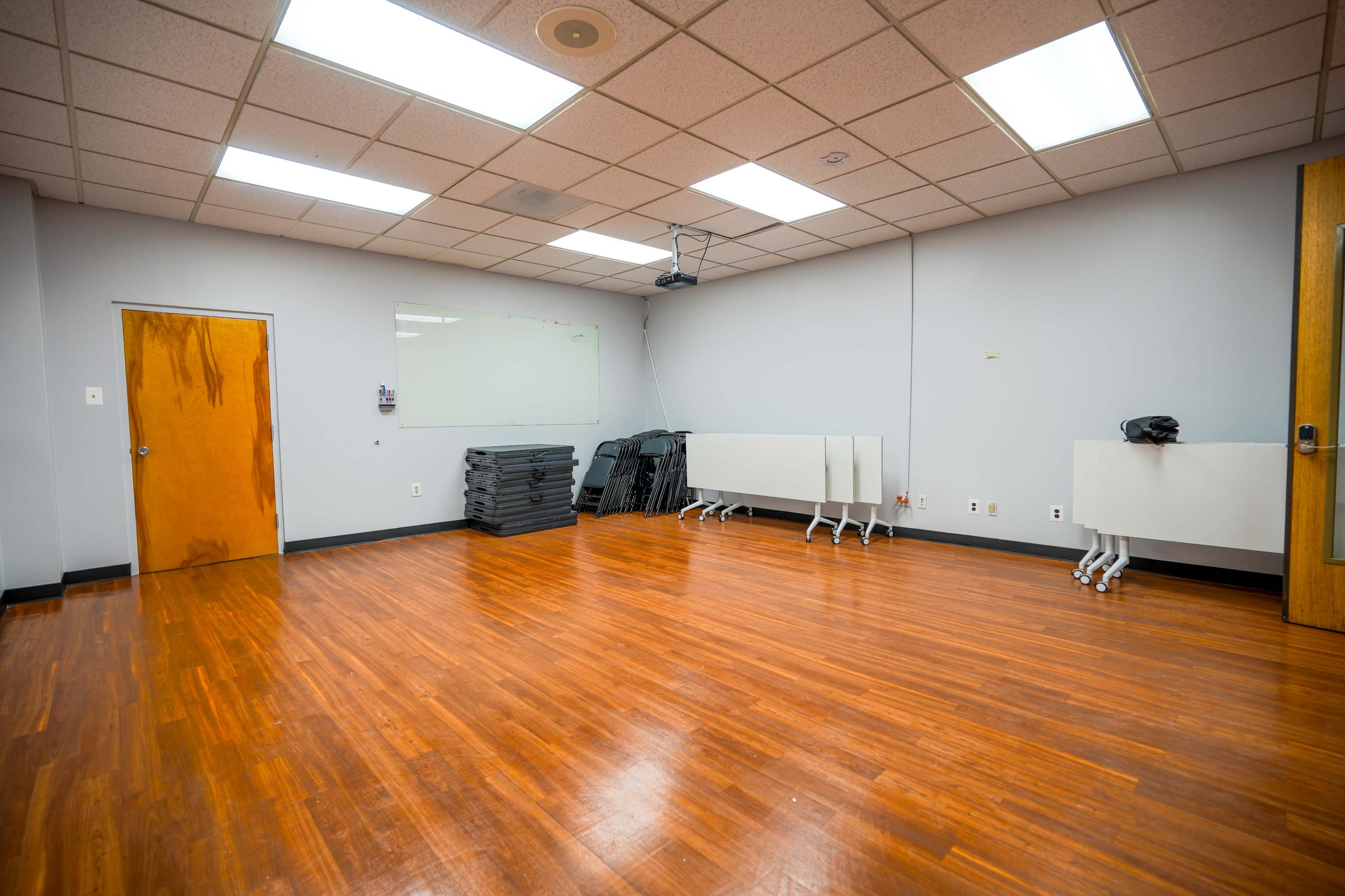 The image shows a large, empty room with polished wooden floors, a wall-mounted projector, and several folding tables stacked against one wall.