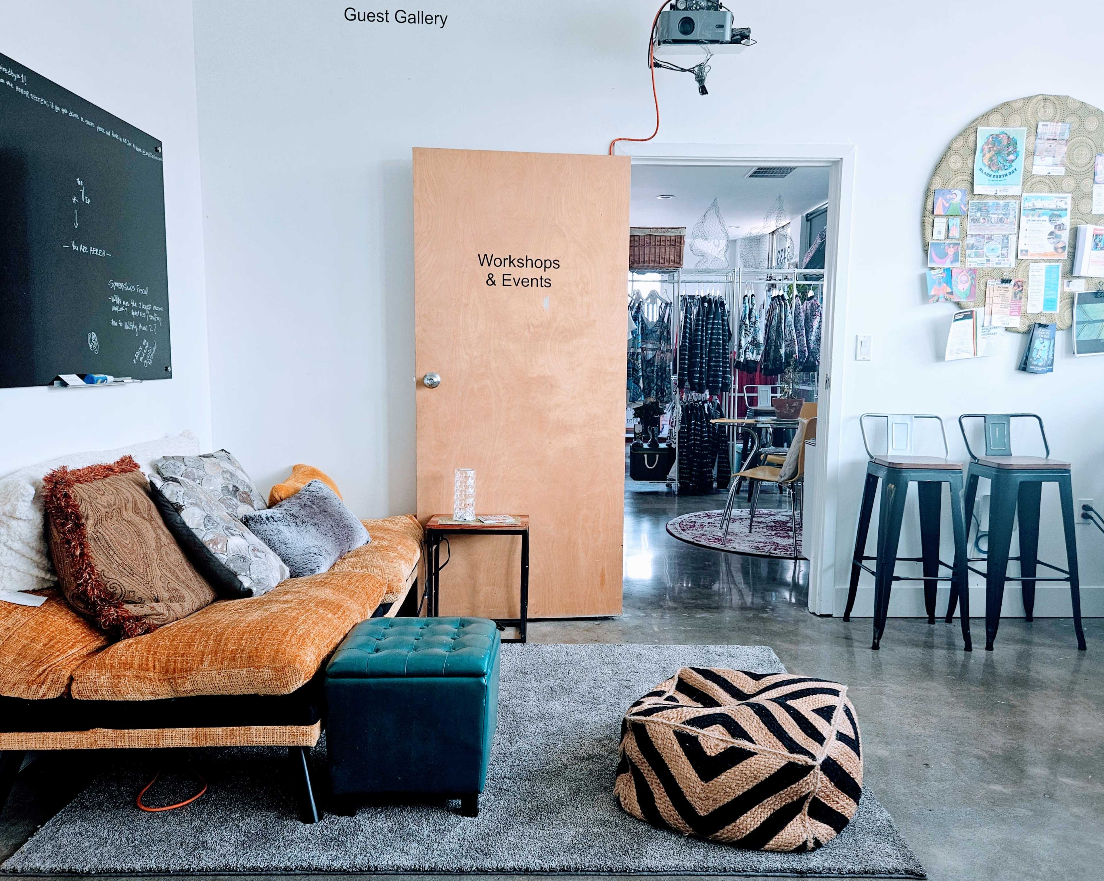 A cozy interior space featuring a sofa with multiple cushions, a round pouf, two bar stools, and a door labeled "Workshops & Events" leading to a gallery area.