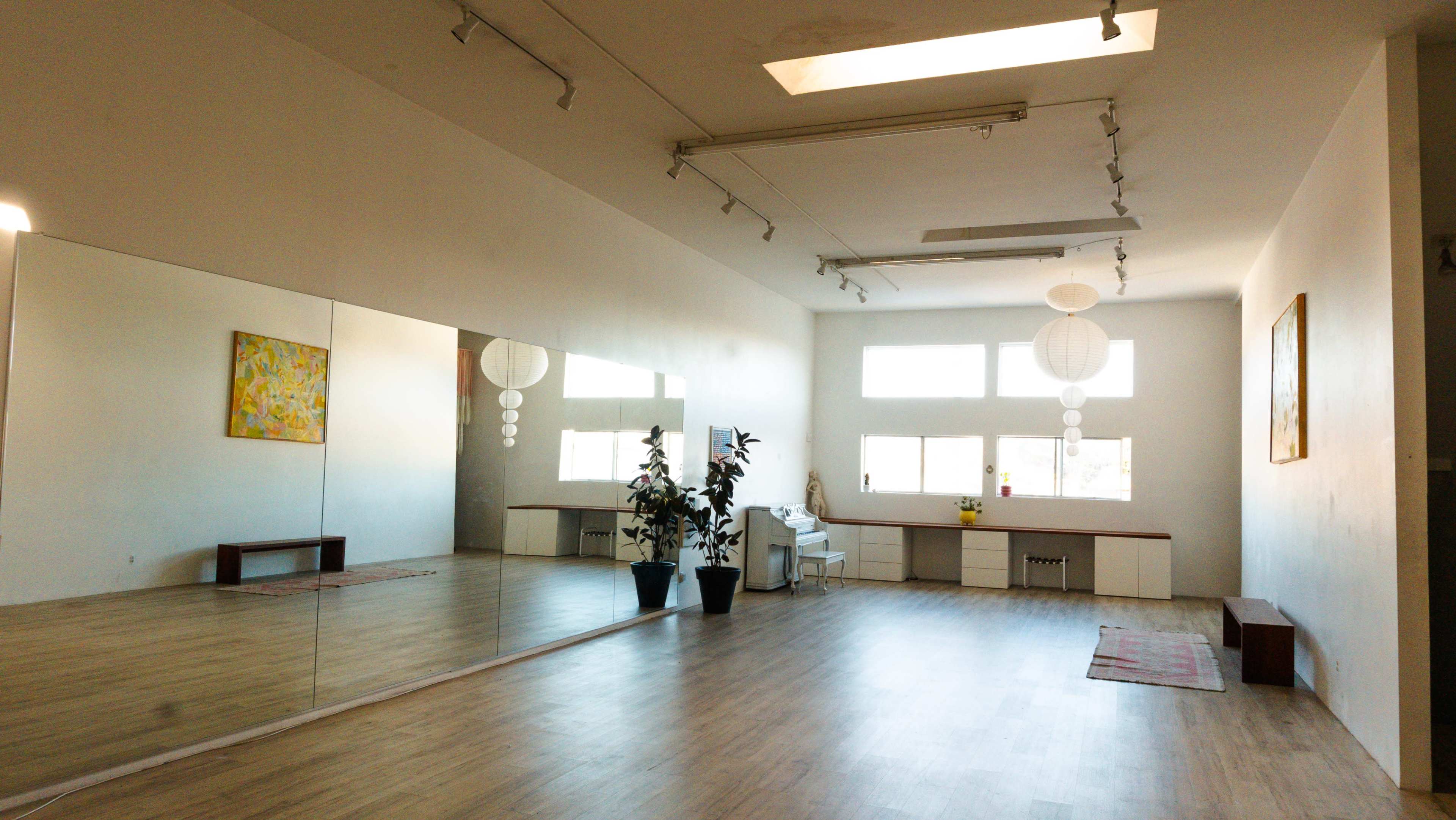 The image shows a spacious, well-lit room with a large mirror on one wall, wooden flooring, and a few decorative items such as a plant and wall art.