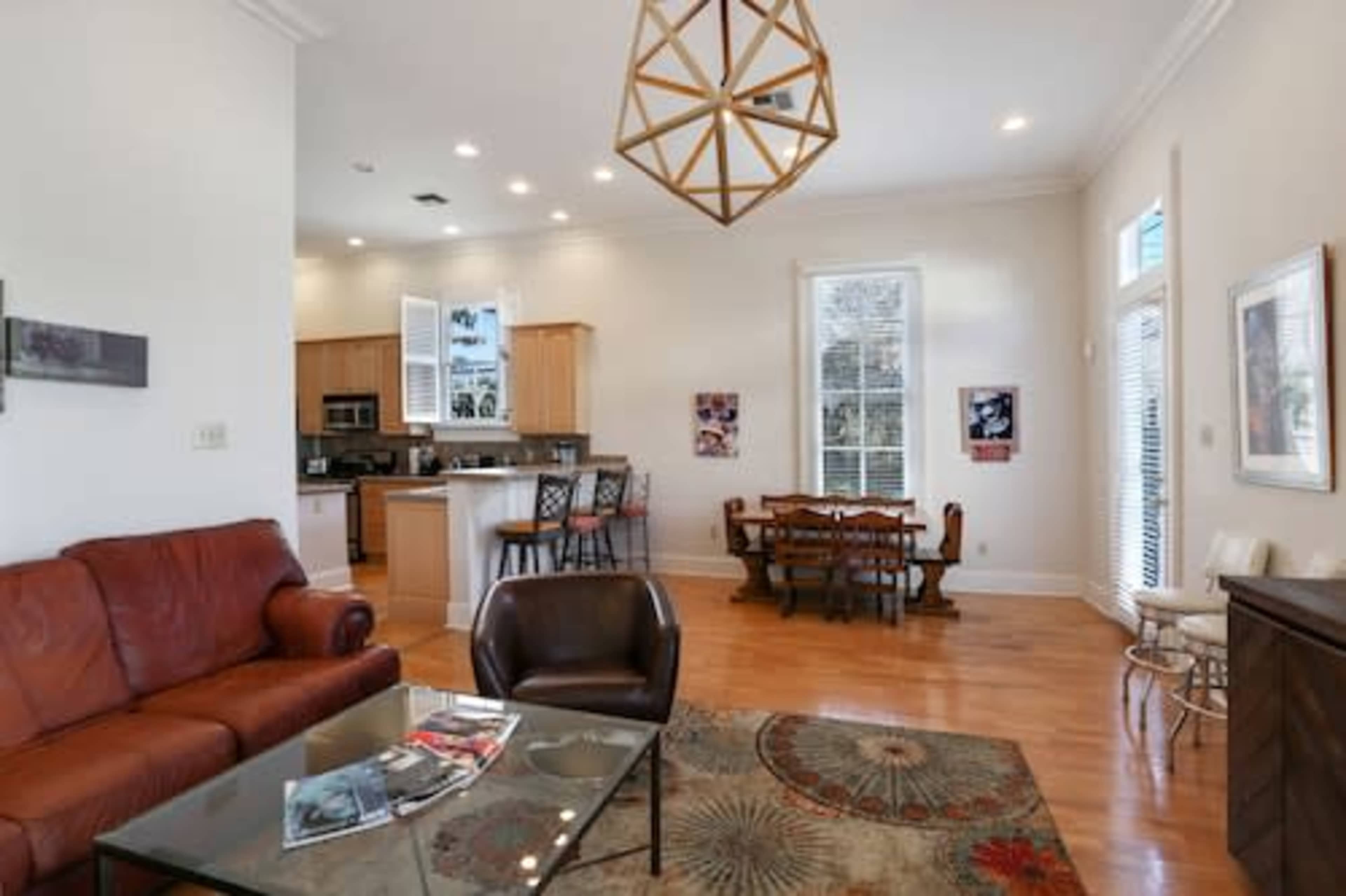 A modern living room and dining area with a leather sofa, coffee table, and a dining table surrounded by chairs, all illuminated by natural light from large windows.