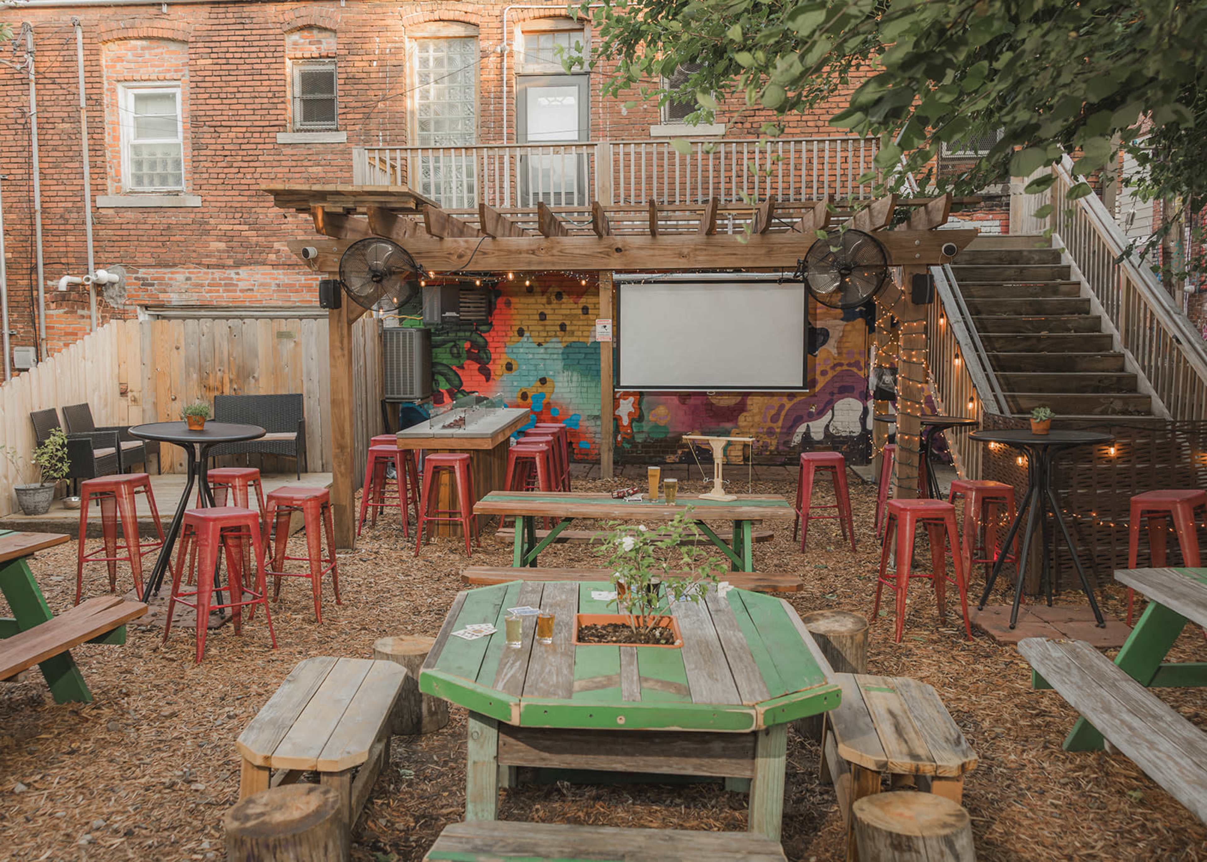 The image shows an outdoor seating area with colorful tables and chairs, surrounded by a mural and a large screen mounted on a wooden structure.