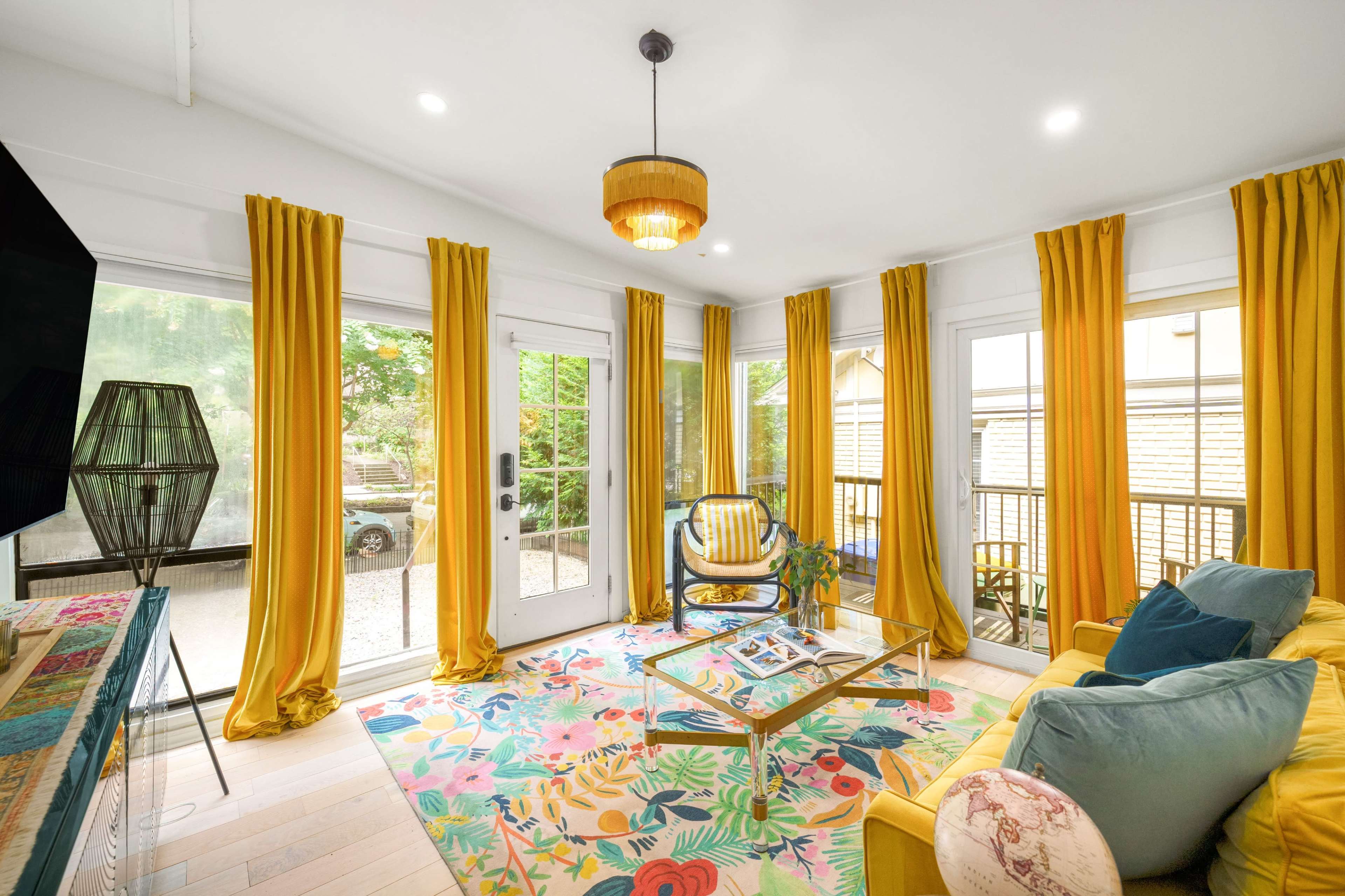 The room features large windows adorned with yellow curtains, a colorful rug, a glass coffee table, and yellow furniture, creating a bright and inviting atmosphere.