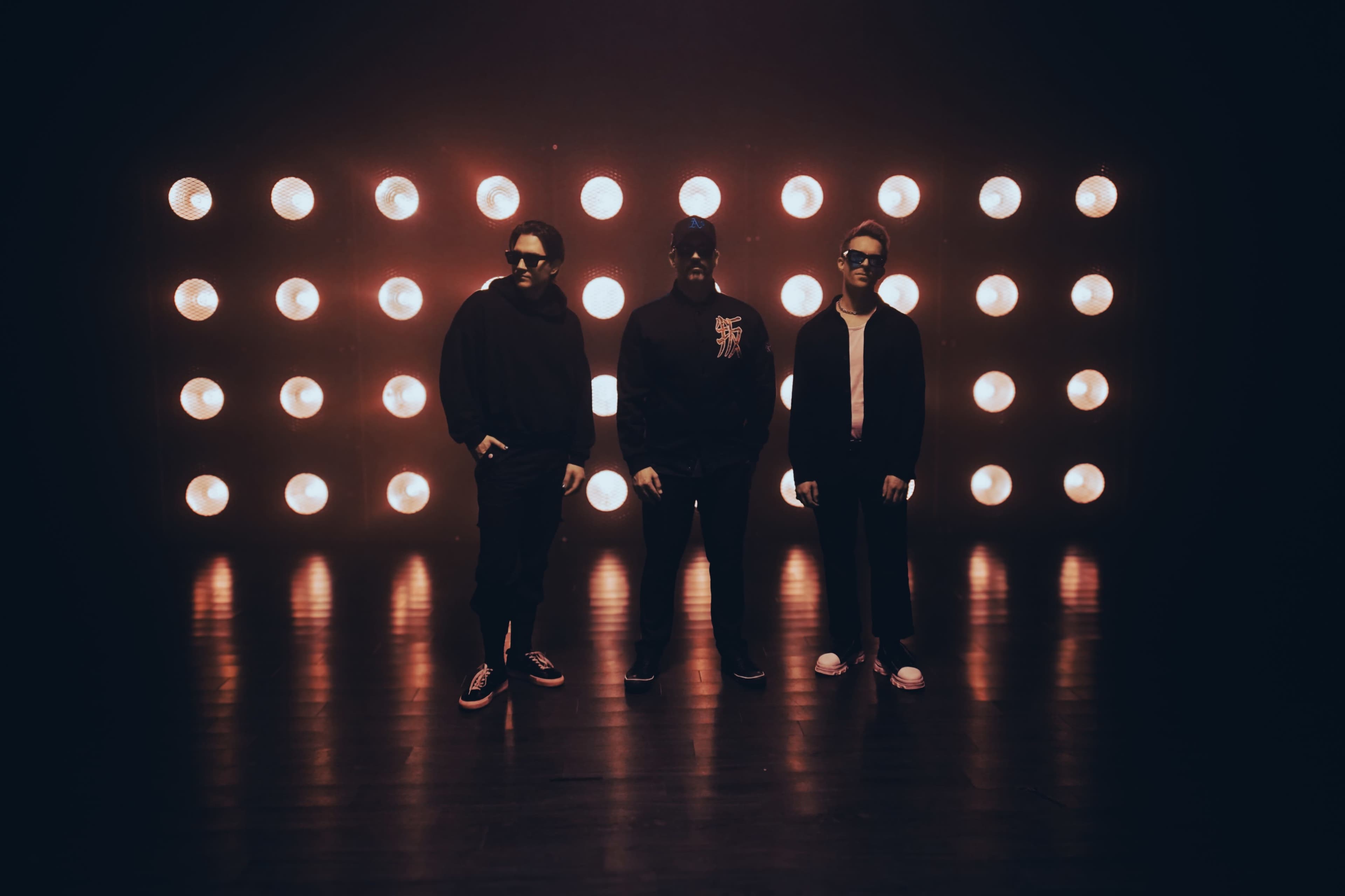 Three individuals stand confidently in front of a wall of bright, round lights in a dimly lit setting.