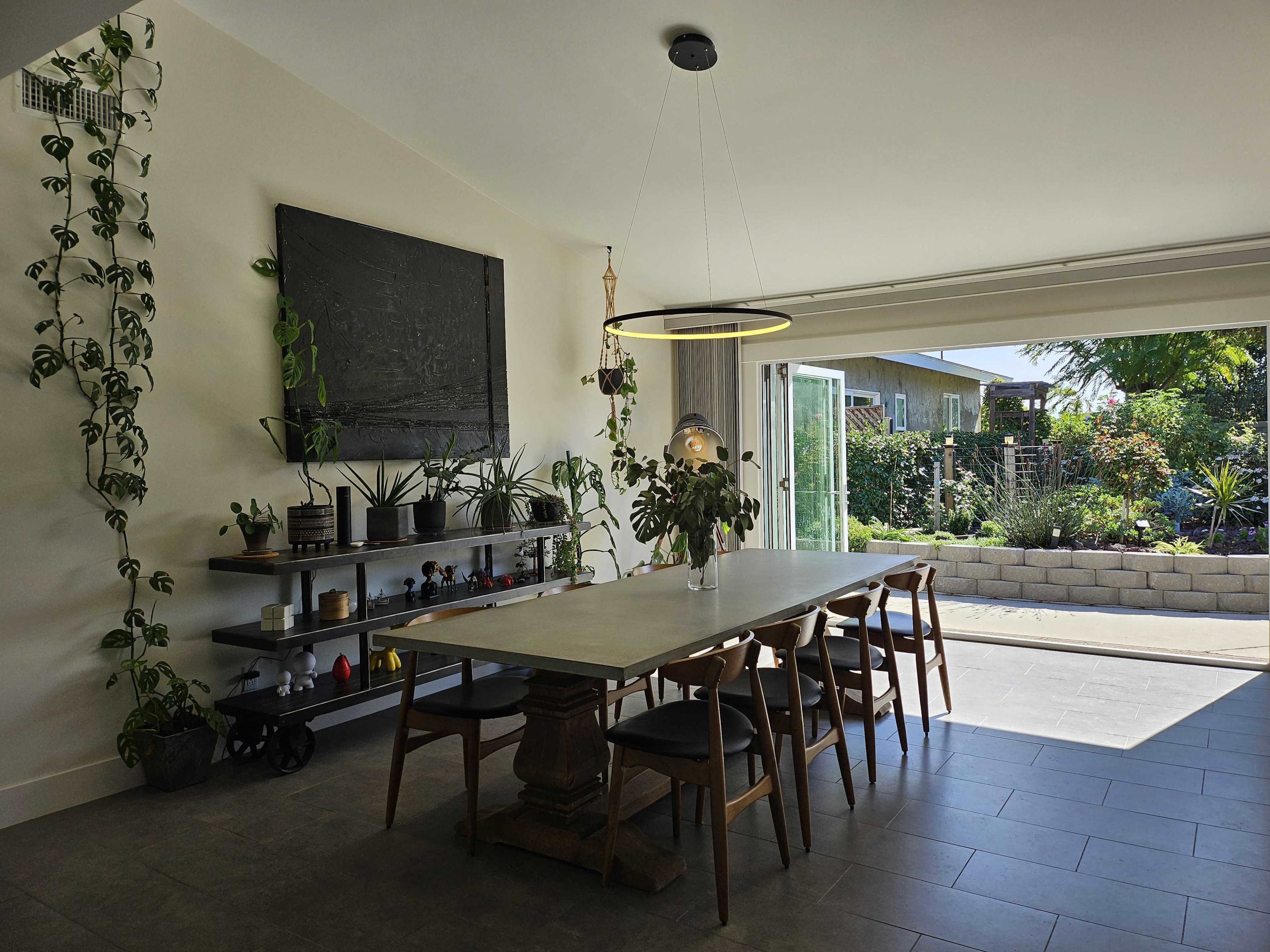 A modern dining area with a large table, surrounded by wooden chairs, and a wall adorned with plants and artwork, leading to a garden space through an open sliding door.
