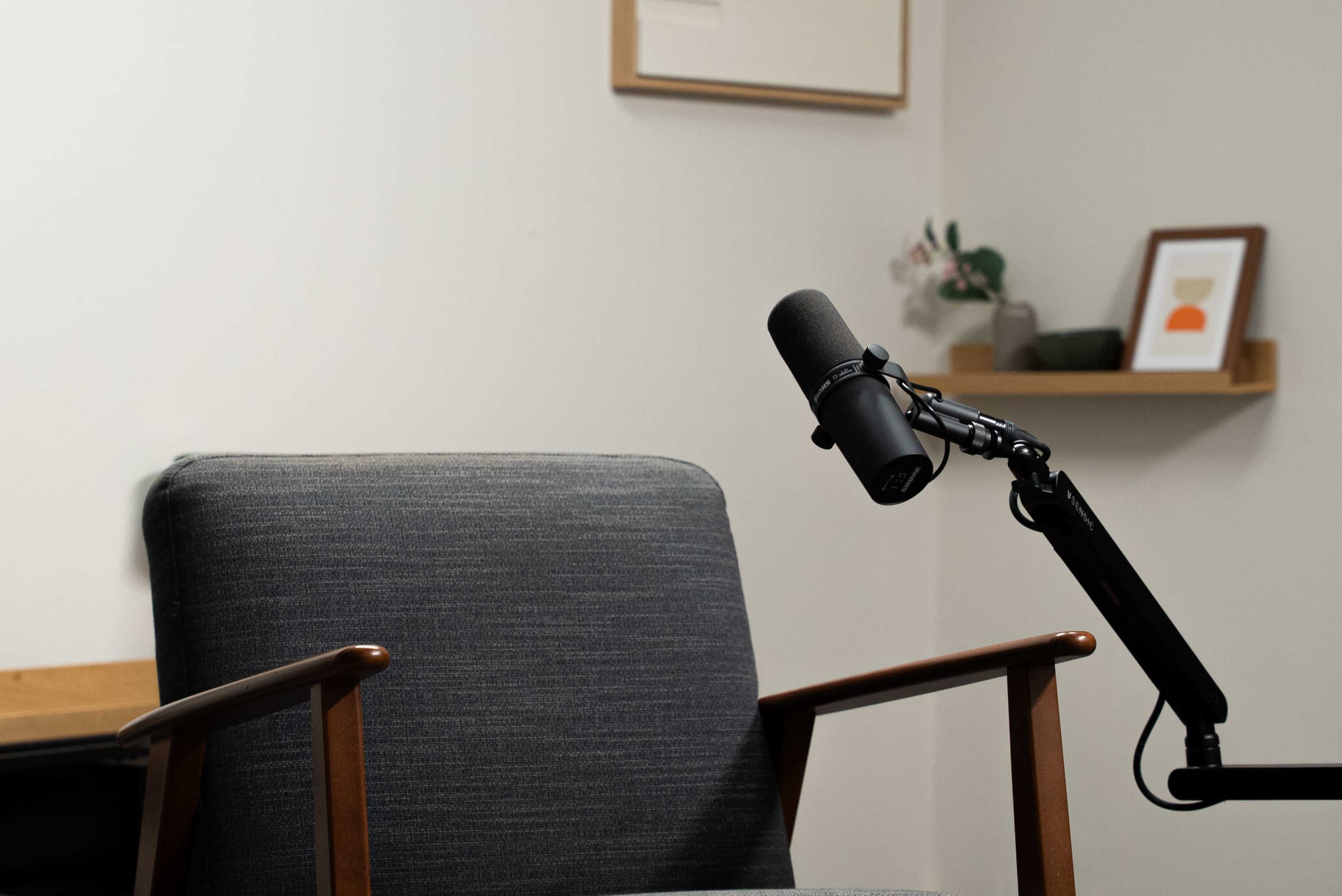 A microphone is positioned beside a gray upholstered chair in a minimalist room with a wall shelf featuring framed art and a small plant.