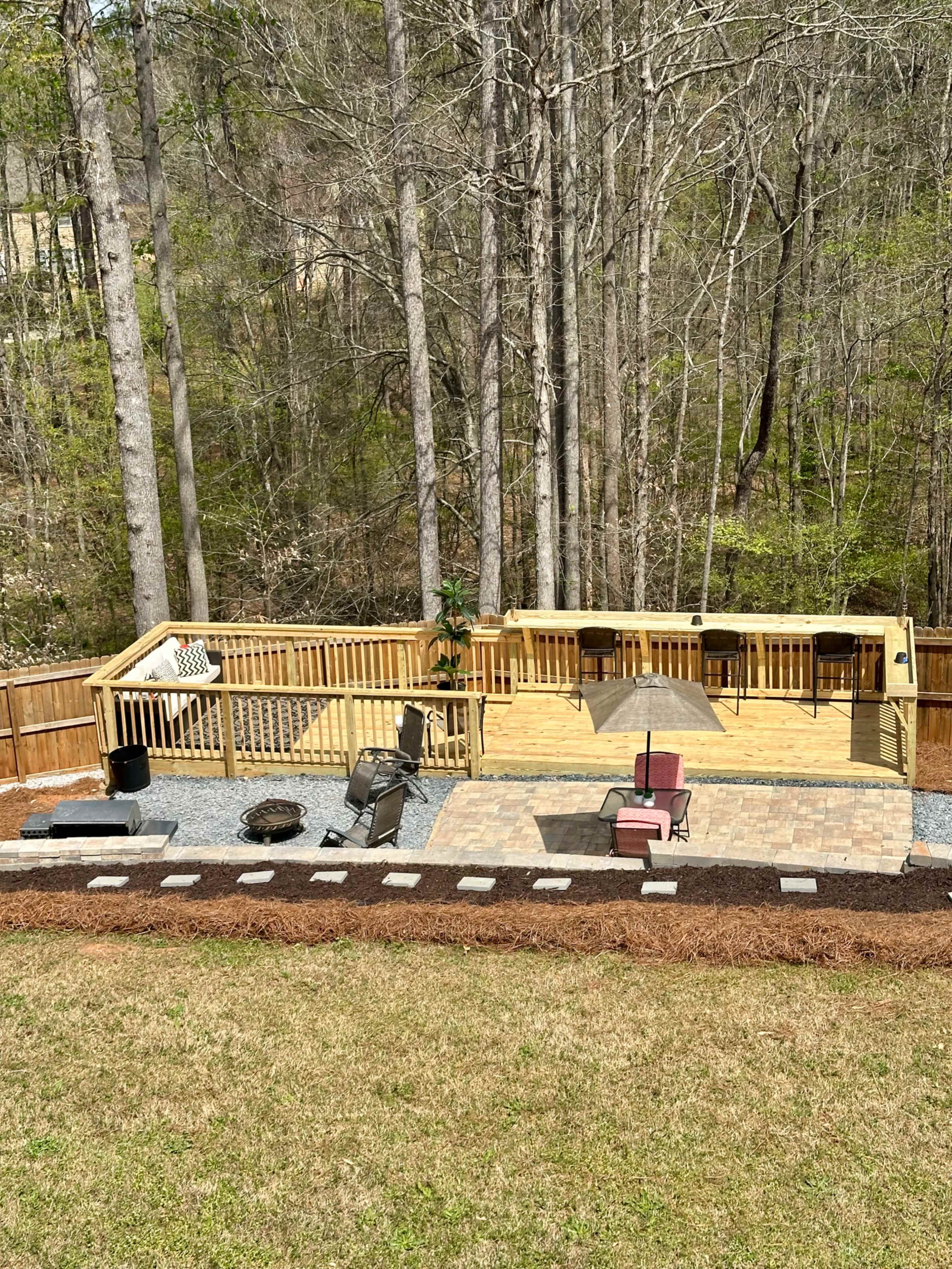 The image shows a wooden deck with patio furniture, a fire pit, and landscaping features, surrounded by trees in a backyard setting.